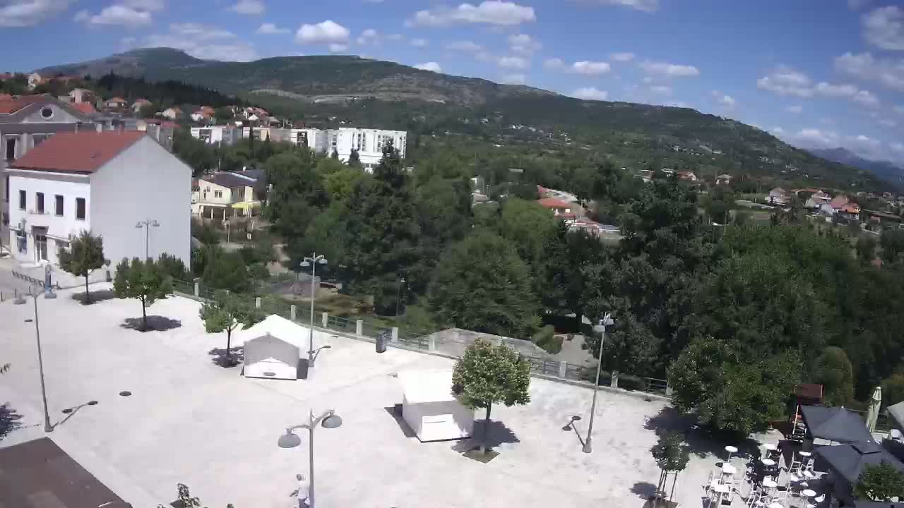 A paved town square with scattered trees and lampposts overlooks a verdant hillside town nestled beneath a mountain range on a partly cloudy day.