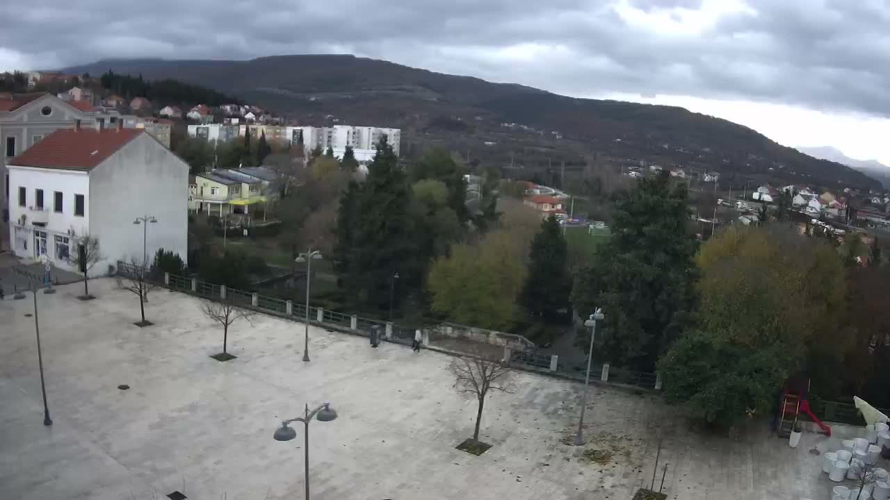 A paved town square with scattered trees and lampposts overlooks a verdant hillside town nestled beneath a mountain range on a partly cloudy day.