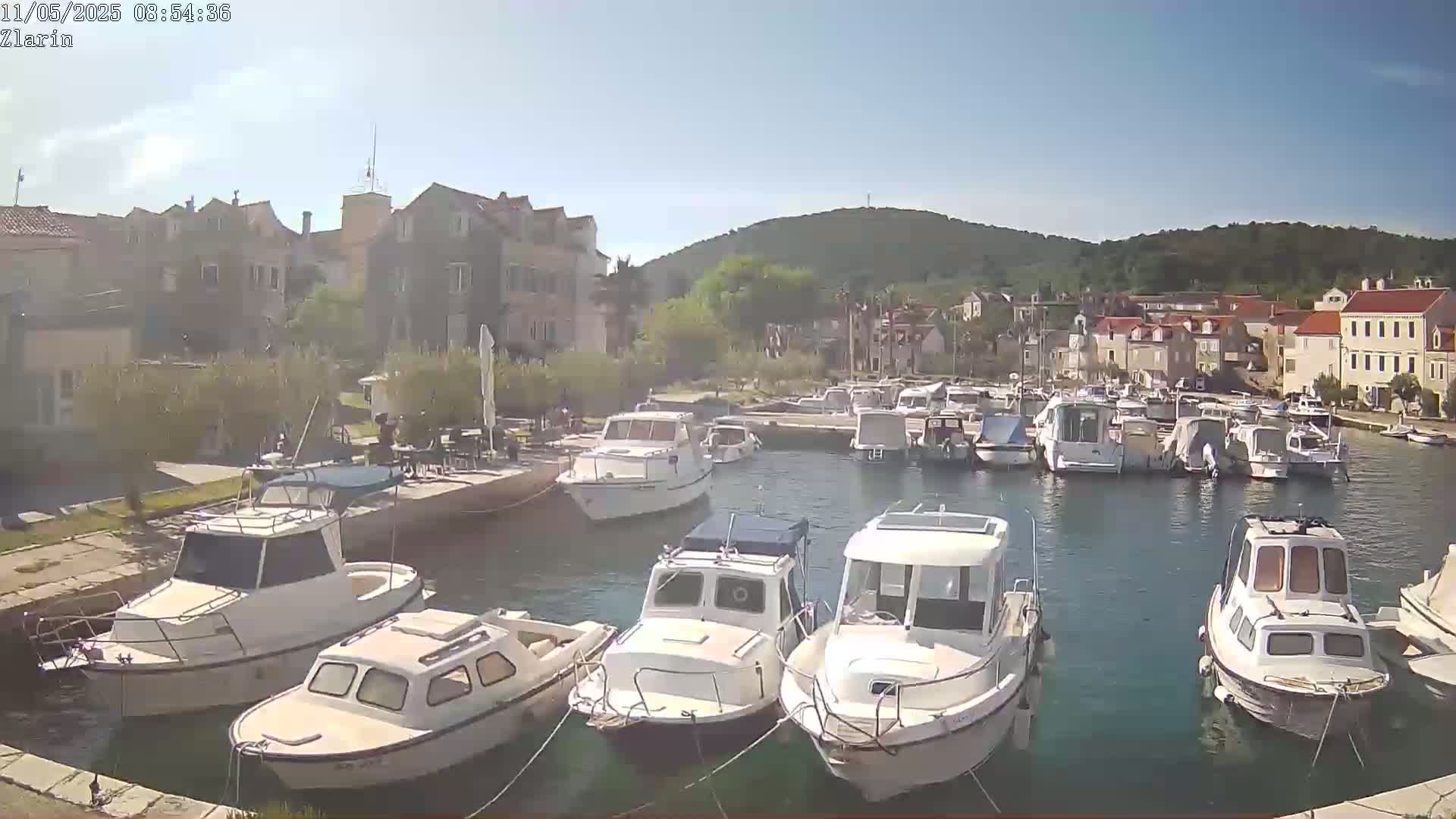 A harbor filled with numerous motorboats is nestled beside a village under a clear, sunny sky, with a hillside visible in the background.