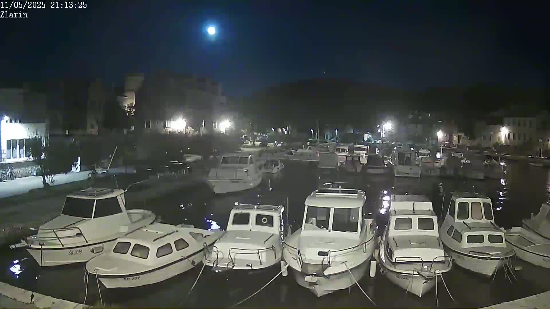 A moonlit night scene shows numerous white motorboats moored in a calm harbor next to a waterfront village.