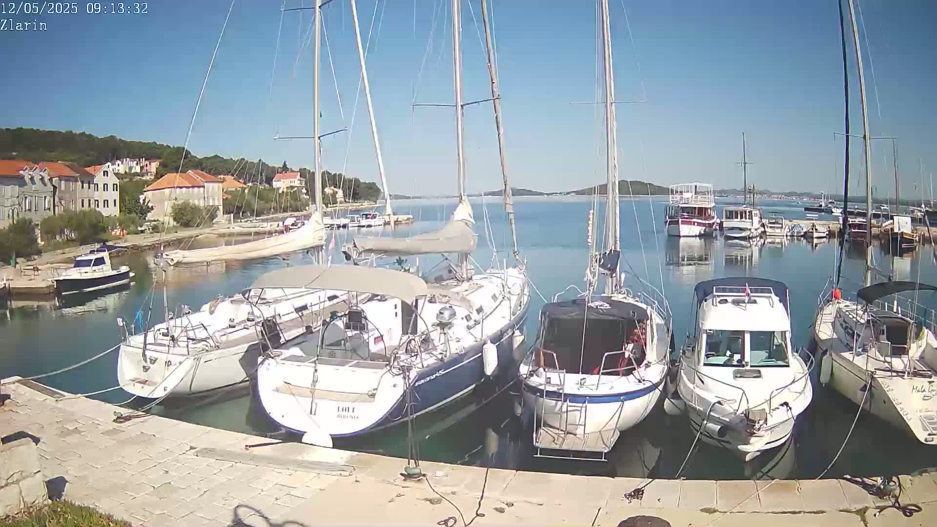 Several sailboats and motorboats are docked in a harbor on a sunny day, with buildings and hills visible in the background.