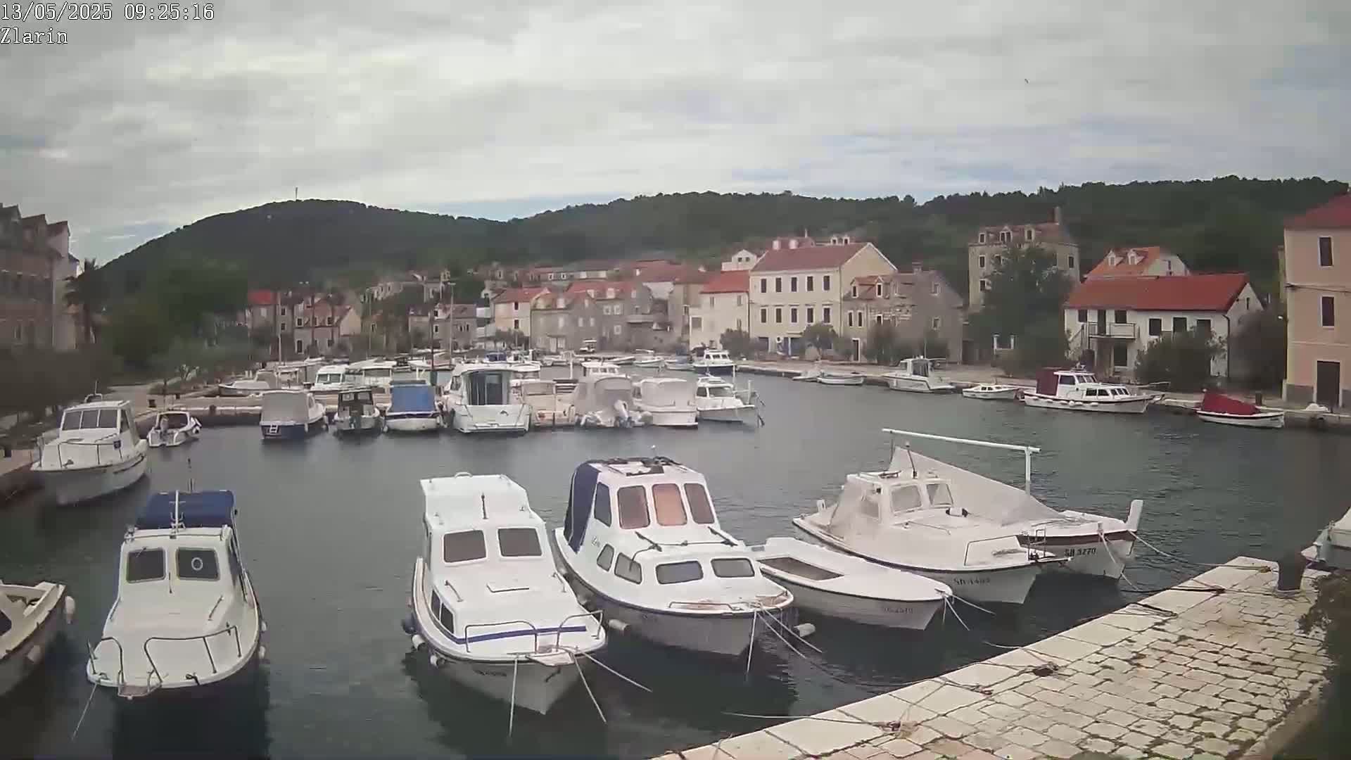 A harbor filled with numerous white motorboats is situated in front of a village of stone buildings under an overcast sky.