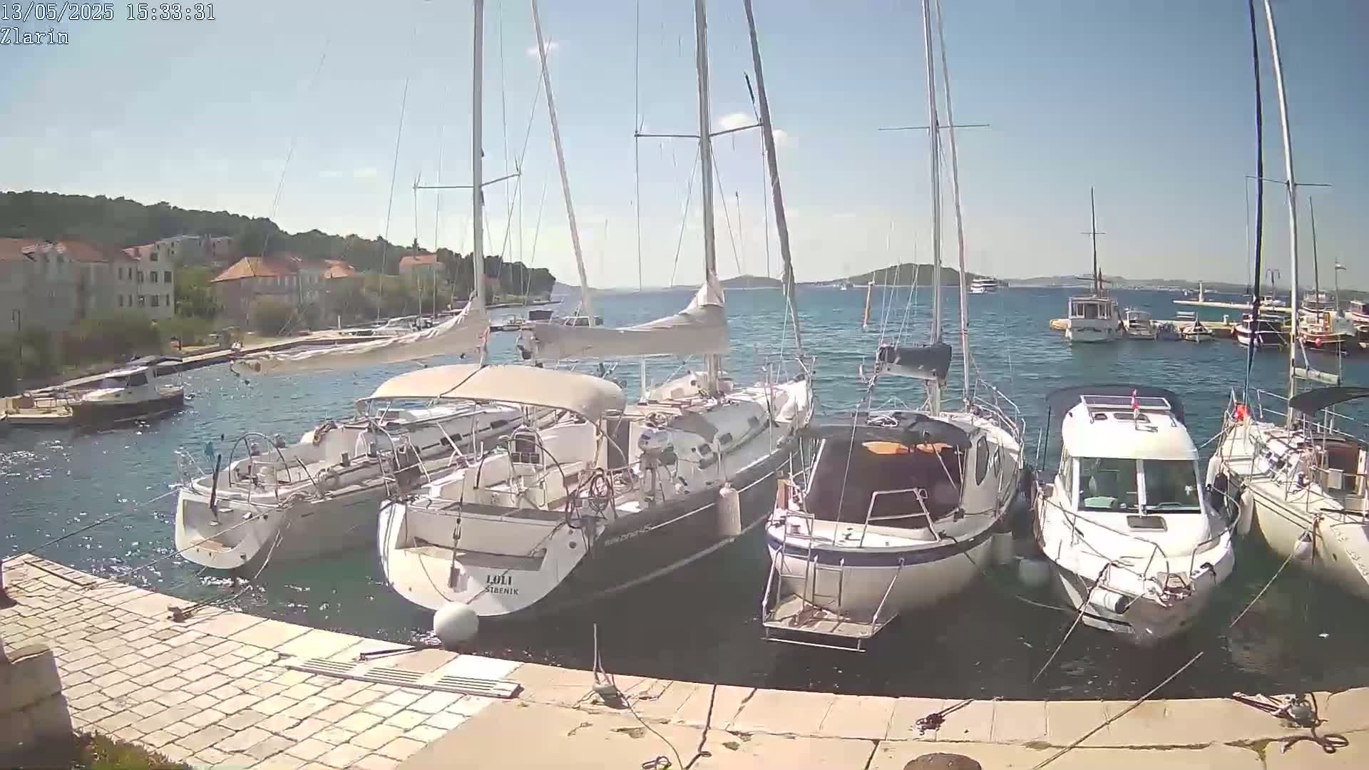 Several sailboats and motorboats are moored in a calm harbor on a sunny day, with a shoreline and buildings visible in the background.