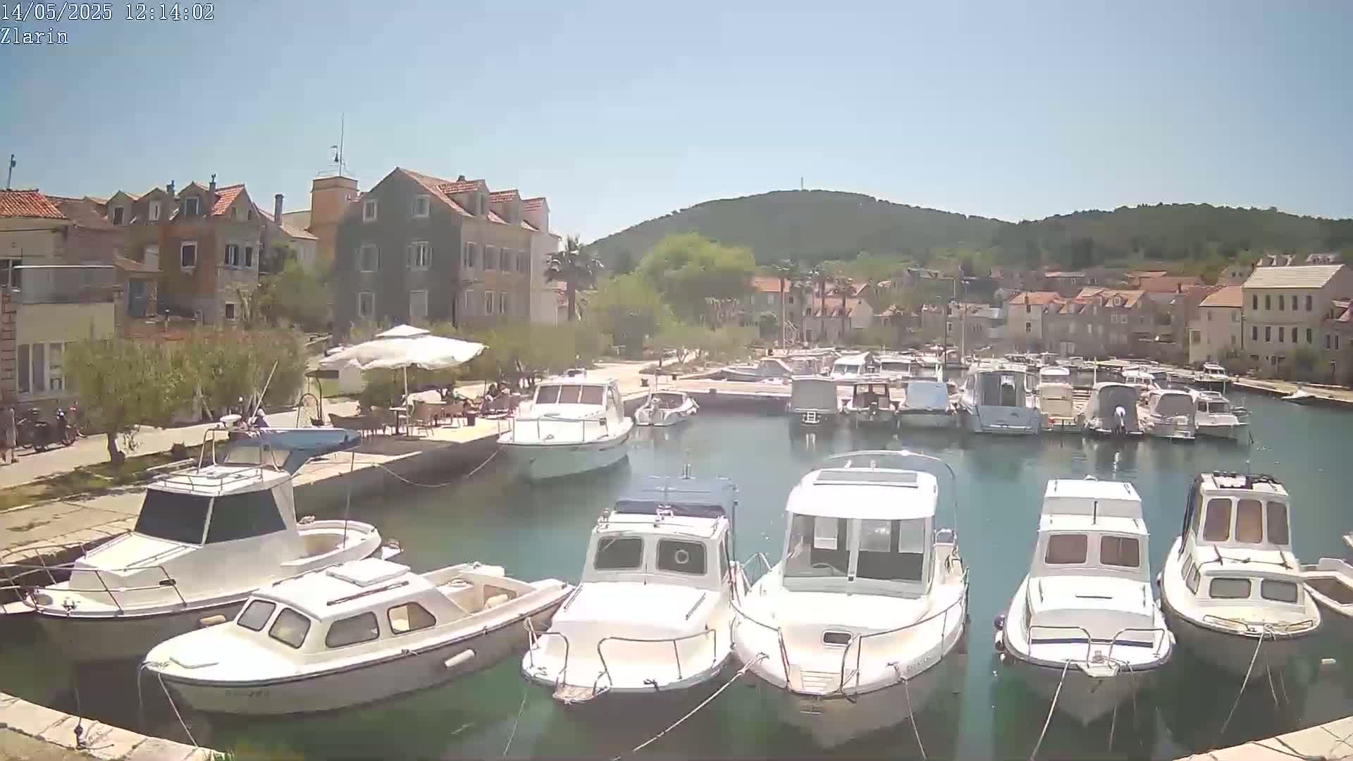 A sunny day shows a harbor filled with numerous white motorboats moored alongside a waterfront lined with buildings and green hills.
