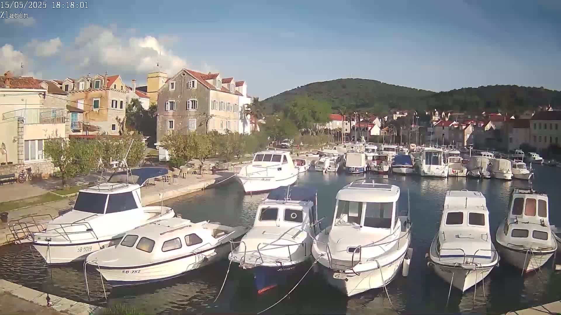 A marina filled with numerous white motorboats is nestled beside a village of stone buildings under a mostly clear, sunny sky.