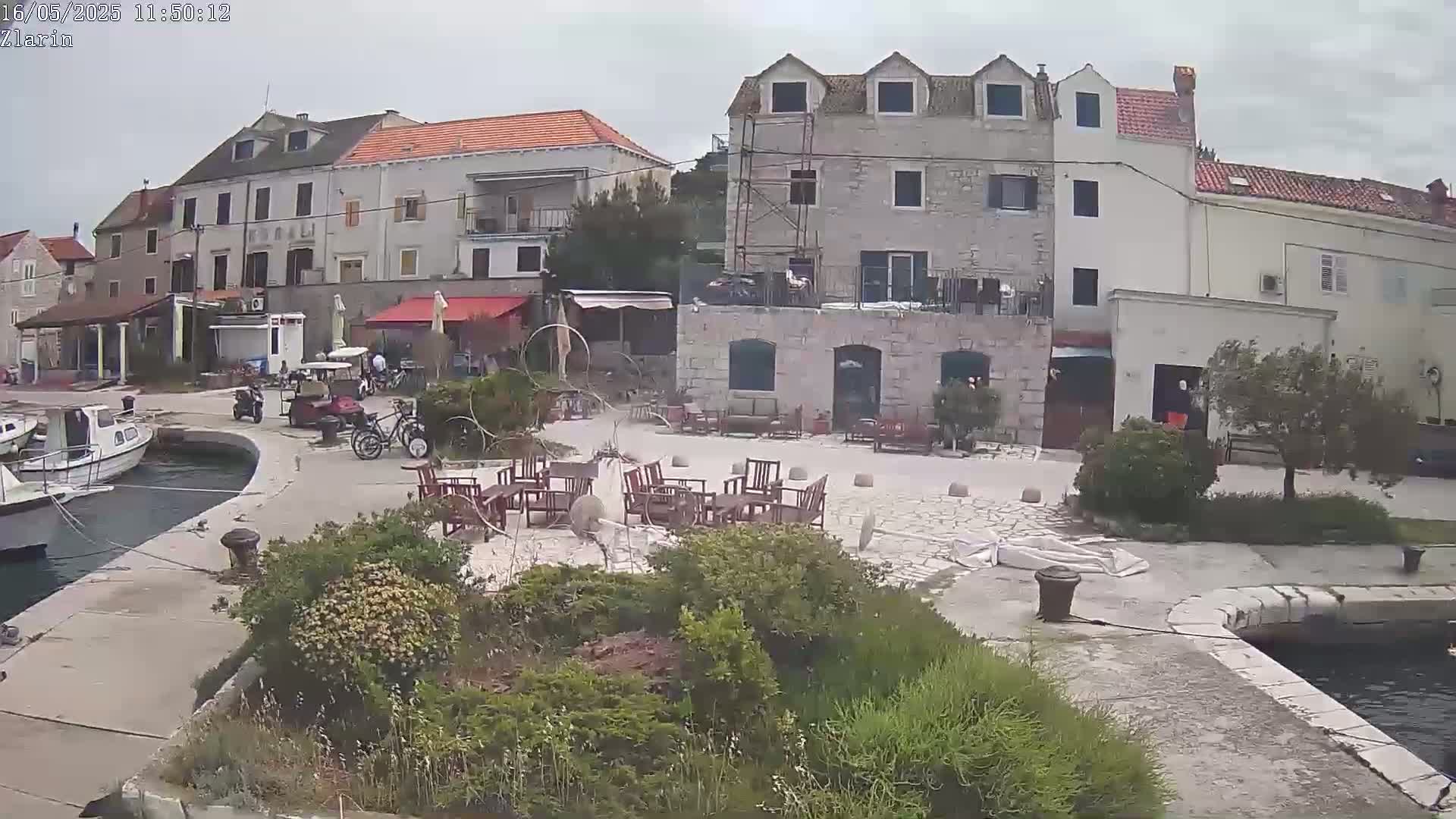 A harborside square with stone buildings, outdoor seating, moored boats, and some overcast weather.