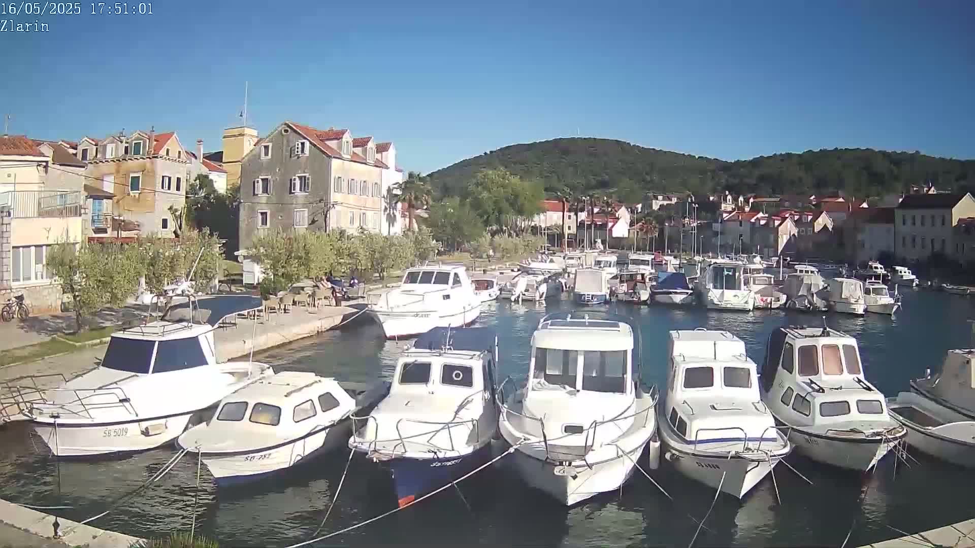 A harbor filled with numerous white motorboats is nestled beside a village of stone buildings under a clear blue sky.