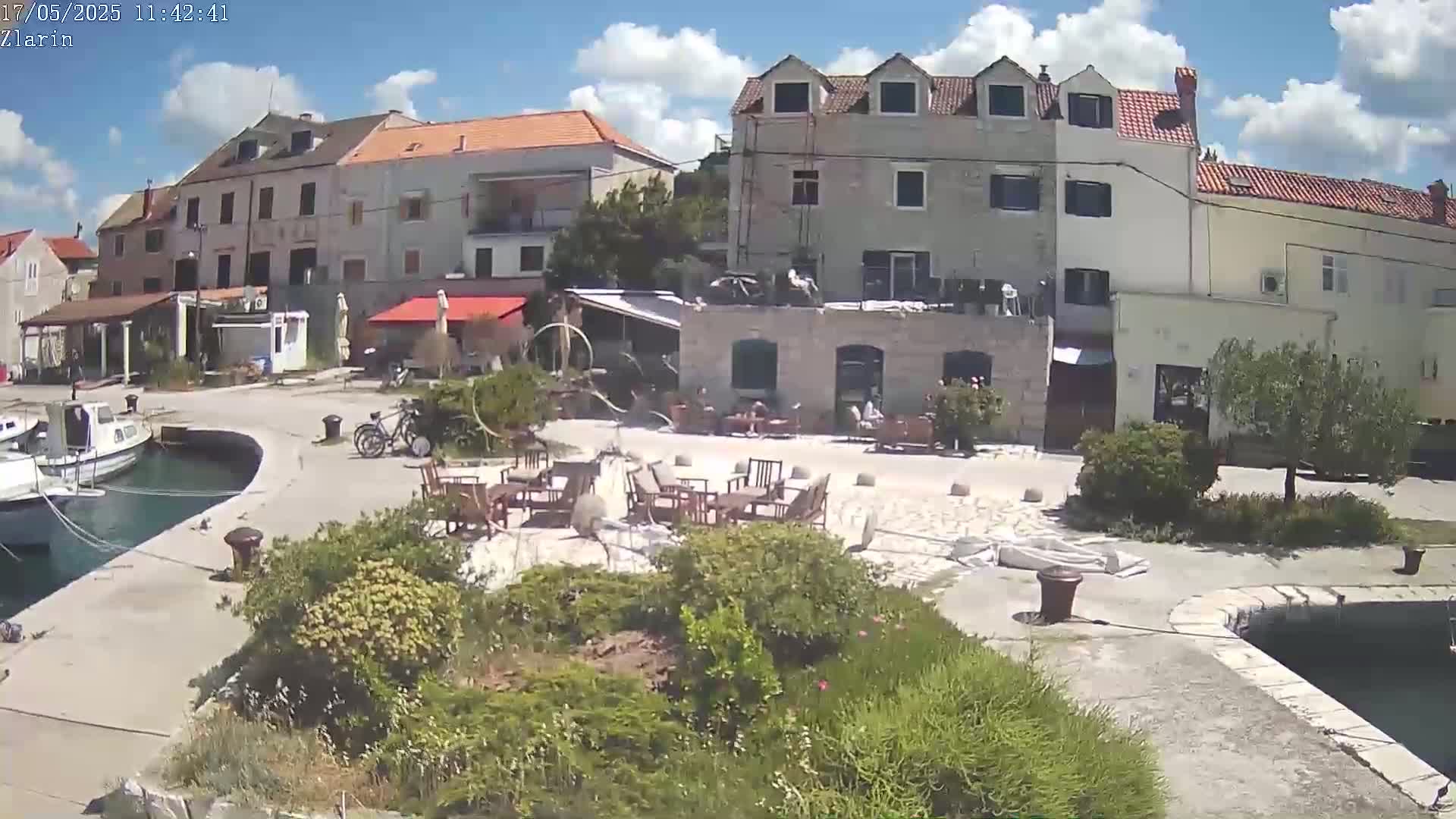 A sunny day shows a waterfront village square with several buildings, outdoor seating, and moored boats.