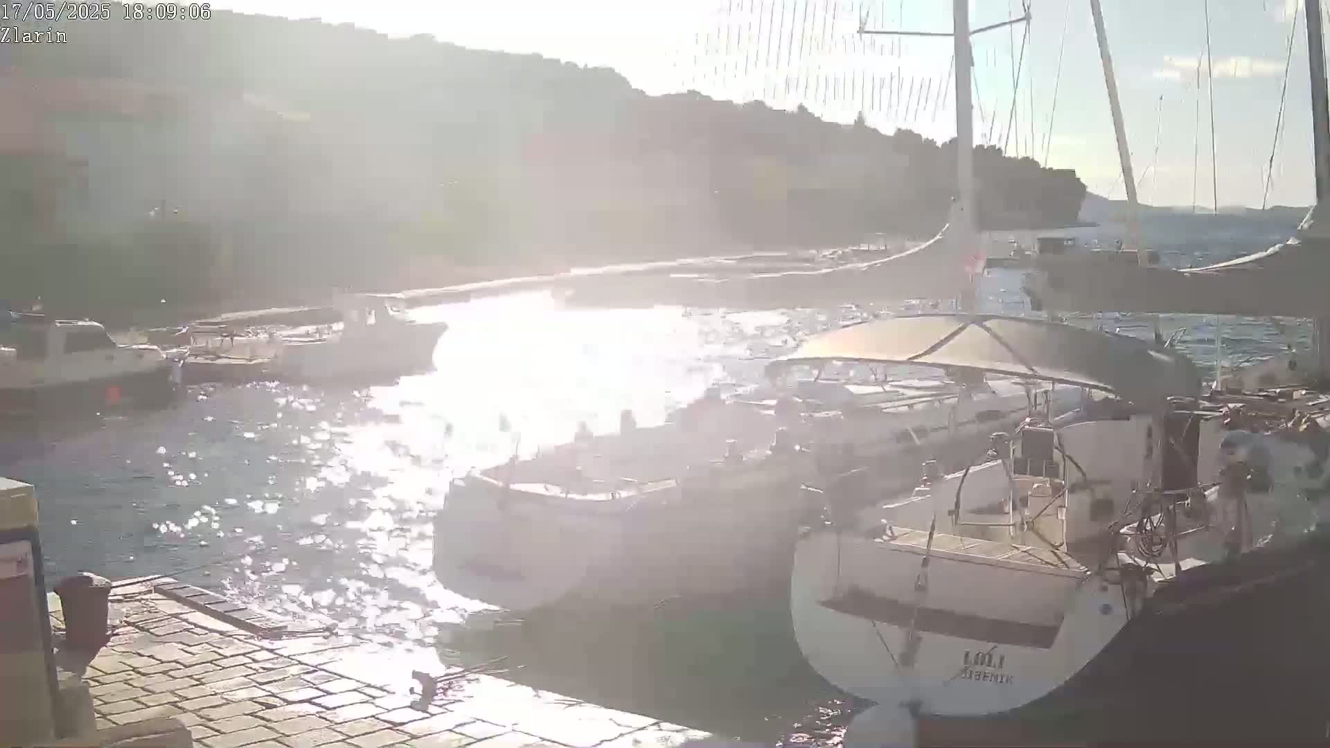 Several boats are moored in a harbor on a sunny day, with sparkling water reflecting the sunlight.
