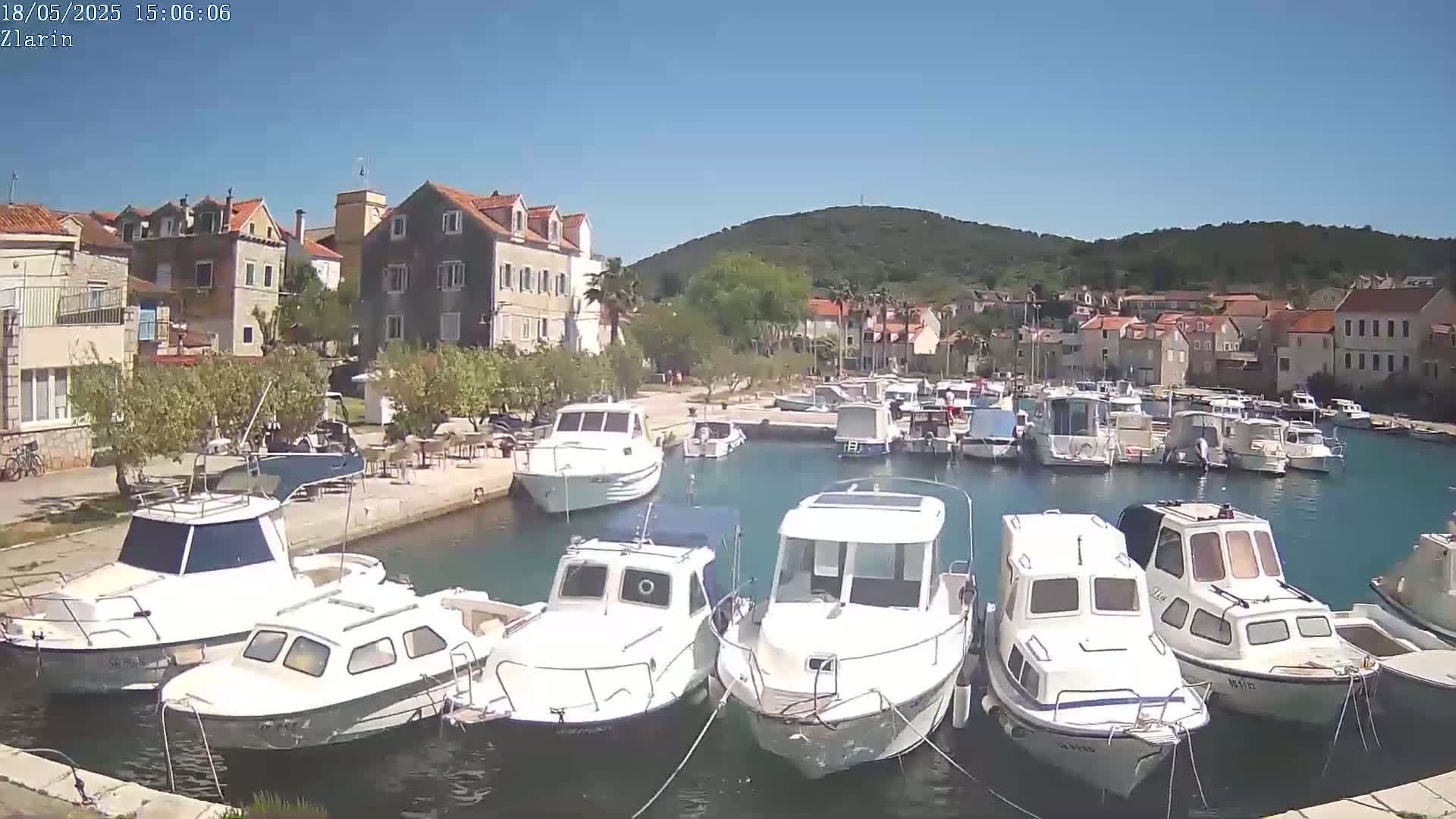 A harbor filled with mostly white motorboats is nestled beside a small village under a clear, sunny sky against a backdrop of a verdant hill.