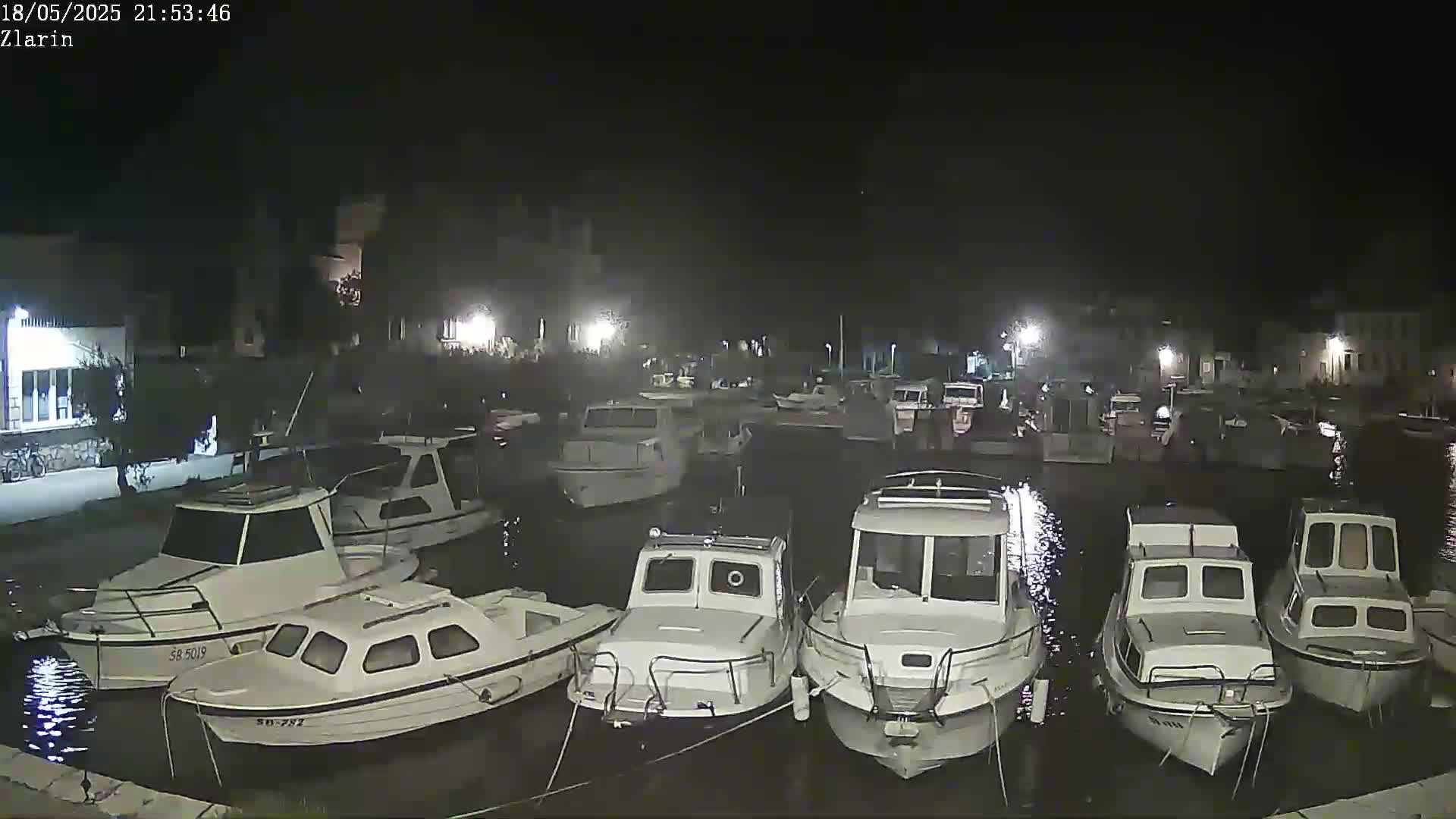 A nighttime harbor scene shows numerous small white boats moored in calm, dark water near a dimly lit shoreline.