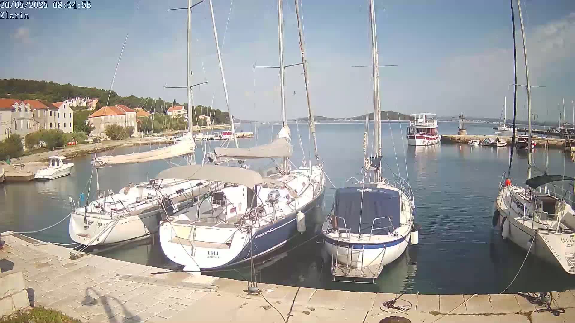 Several sailboats are moored in a calm harbor next to a stone quay, with a small town and surrounding hills visible in the background under a clear, sunny sky.