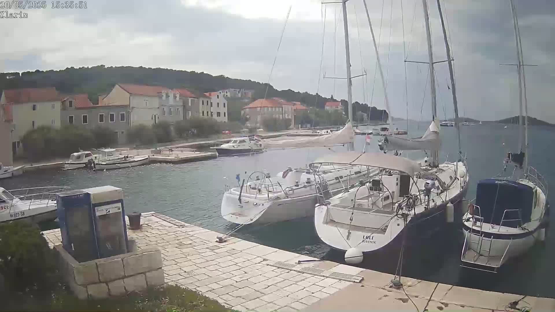 Several sailboats and motorboats are moored at a harbor next to a small town under an overcast sky.