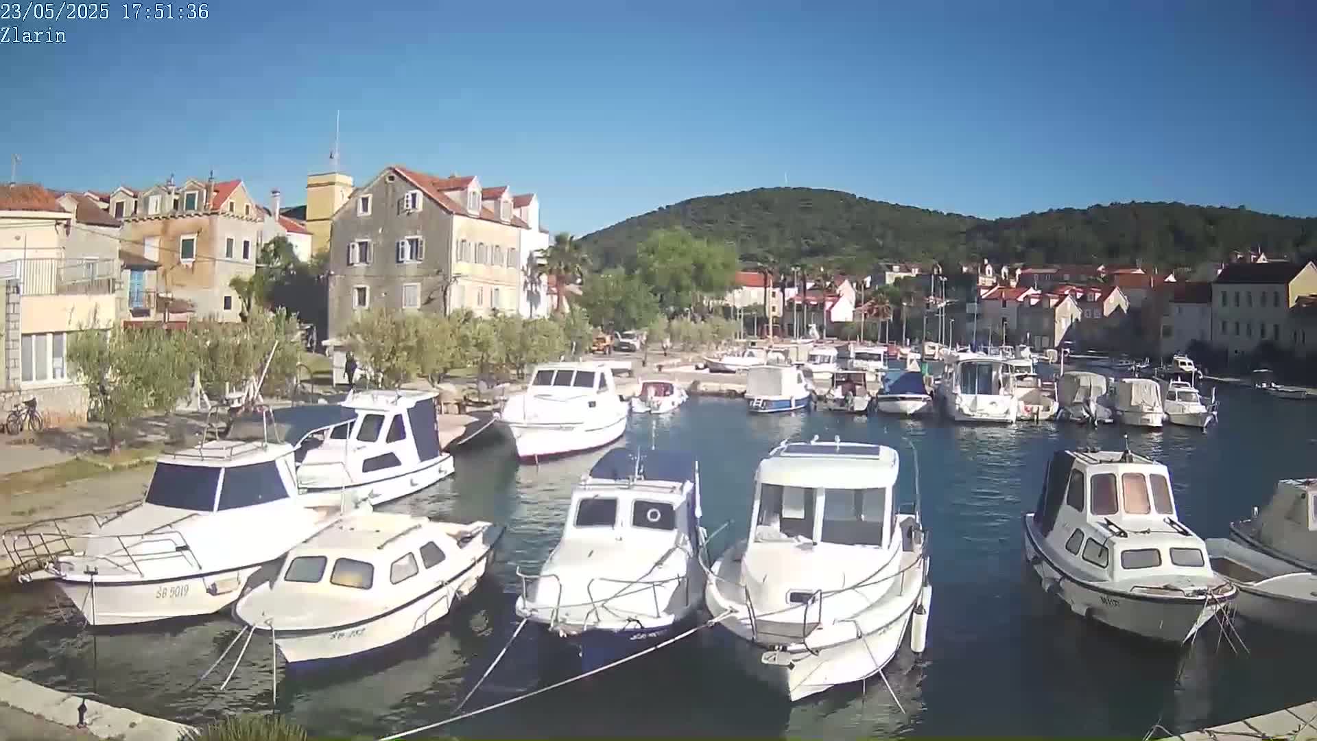 A sunny day in a harbor town shows numerous boats moored in calm water, surrounded by low buildings and a verdant hillside in the background.