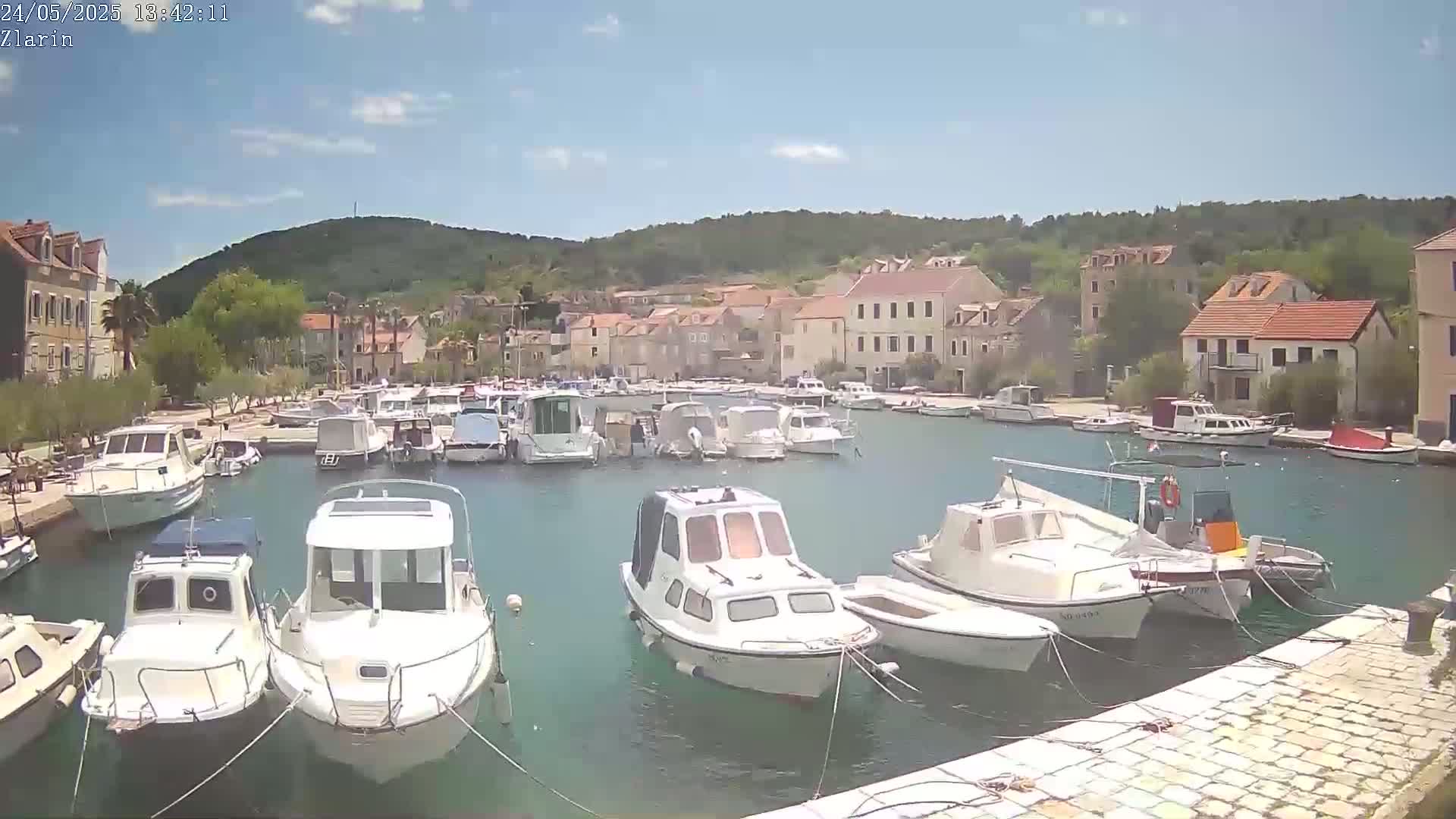 A harbor filled with numerous boats is nestled in a small town on a sunny day, with hills covered in green vegetation in the background.