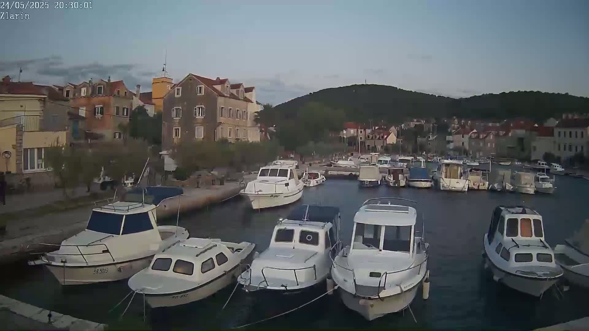 Numerous white motorboats are moored in a calm harbor next to a village of stone buildings, under a dusky sky.
