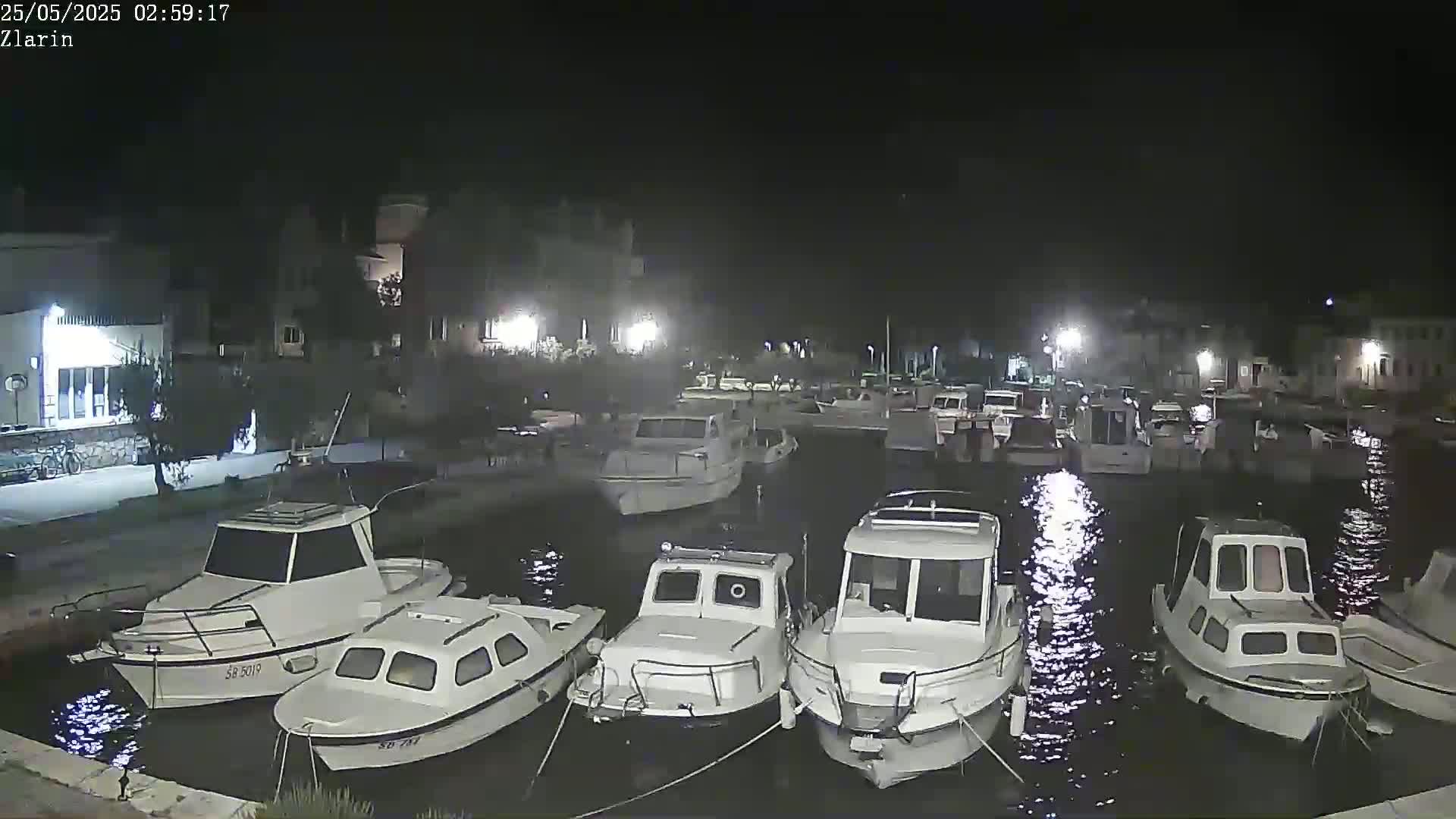 A nighttime harbor scene shows numerous small motorboats moored in calm water, illuminated by lights from nearby buildings.