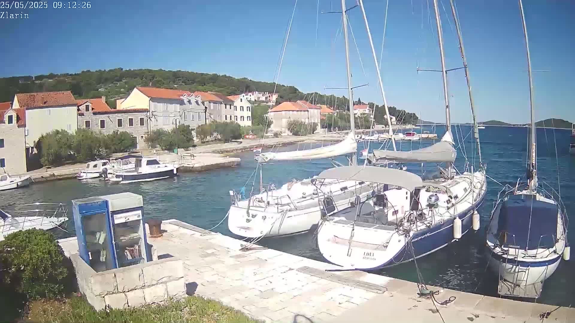 Several sailboats are moored in a calm harbor next to a small village under a clear, sunny sky.