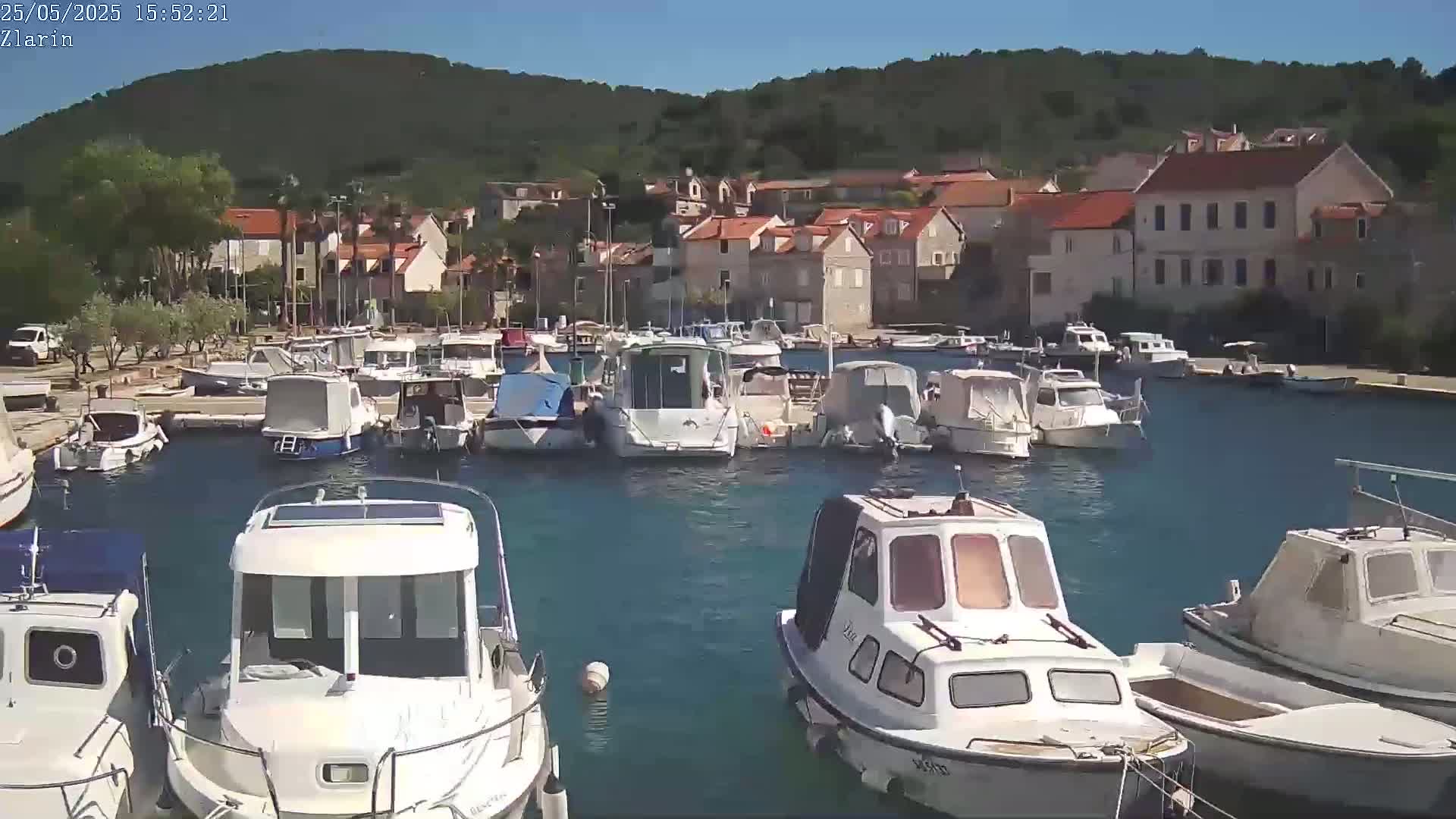 A sunny day shows a harbor filled with numerous small motorboats docked near a village of stone buildings nestled against a green hillside.