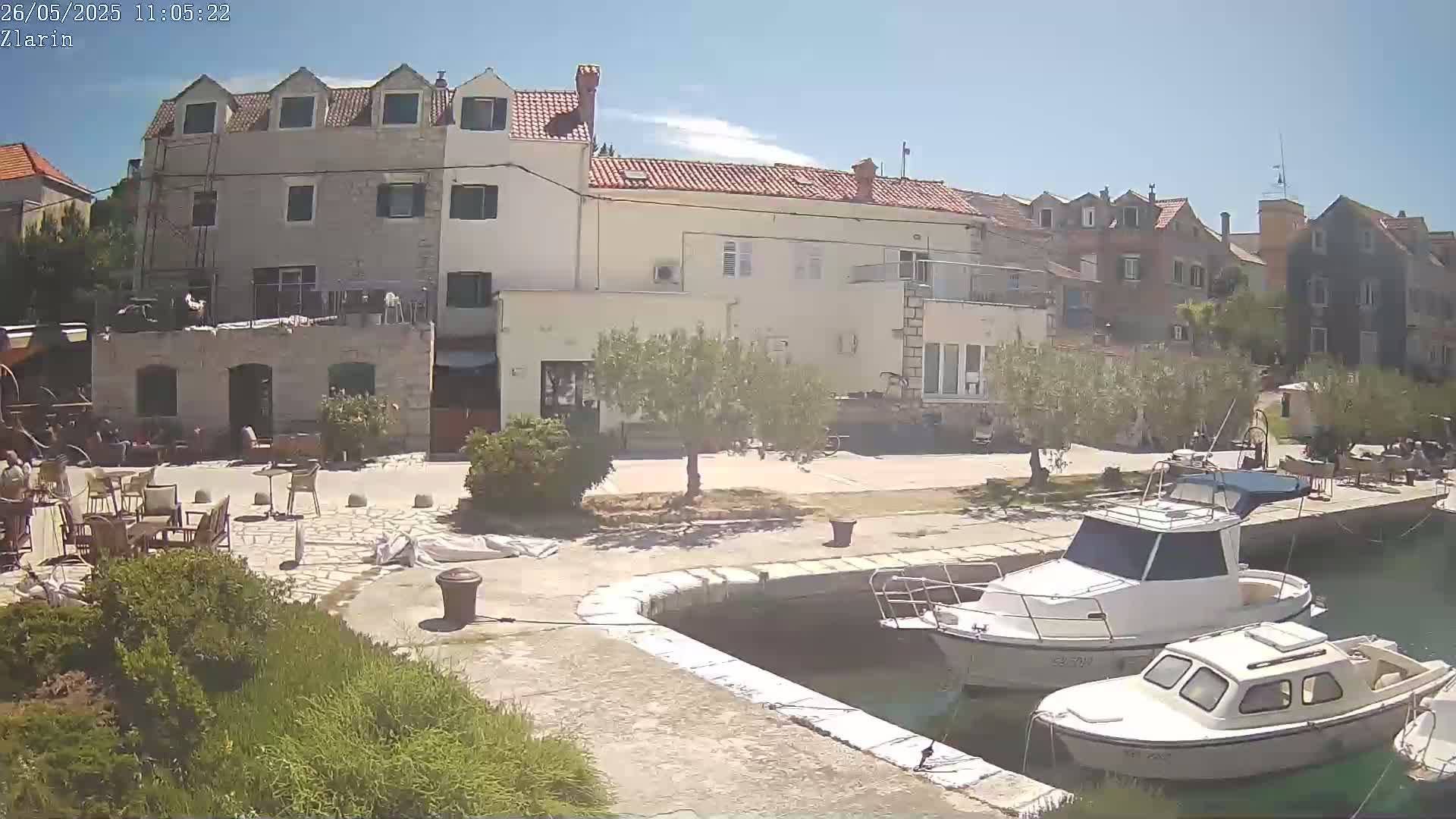 A waterfront scene on a sunny day shows a stone building with outdoor seating, olive trees, and several small boats docked in calm water.