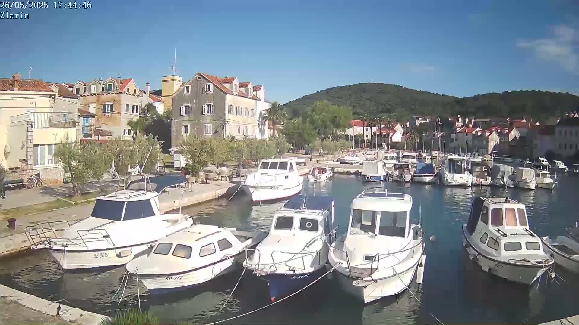 A harbor filled with numerous white motorboats is nestled beside a village of stone buildings under a clear blue sky.