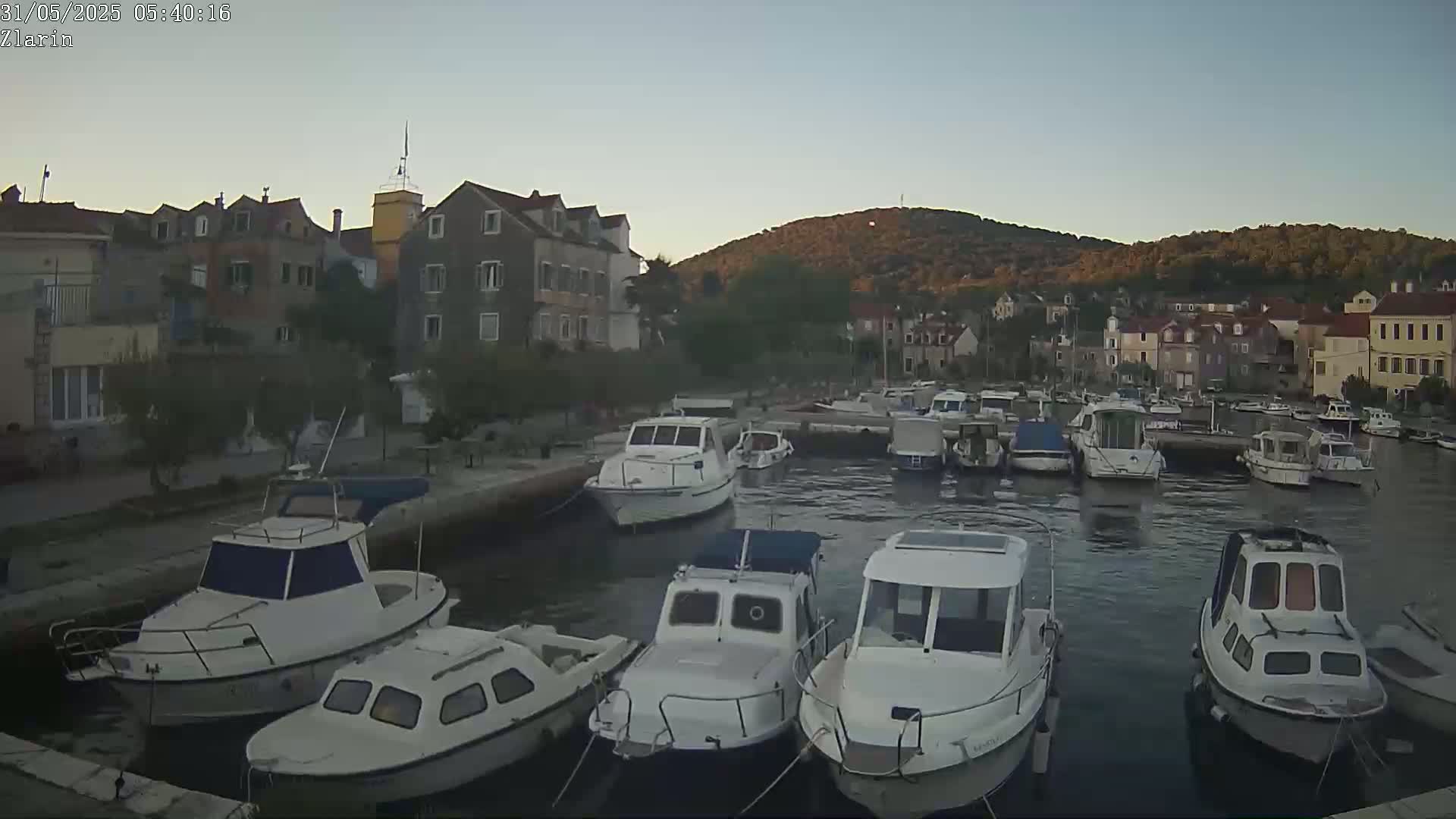 A calm harbor filled with numerous small motorboats is nestled beside a village of stone buildings on a hill, under a clear twilight sky.