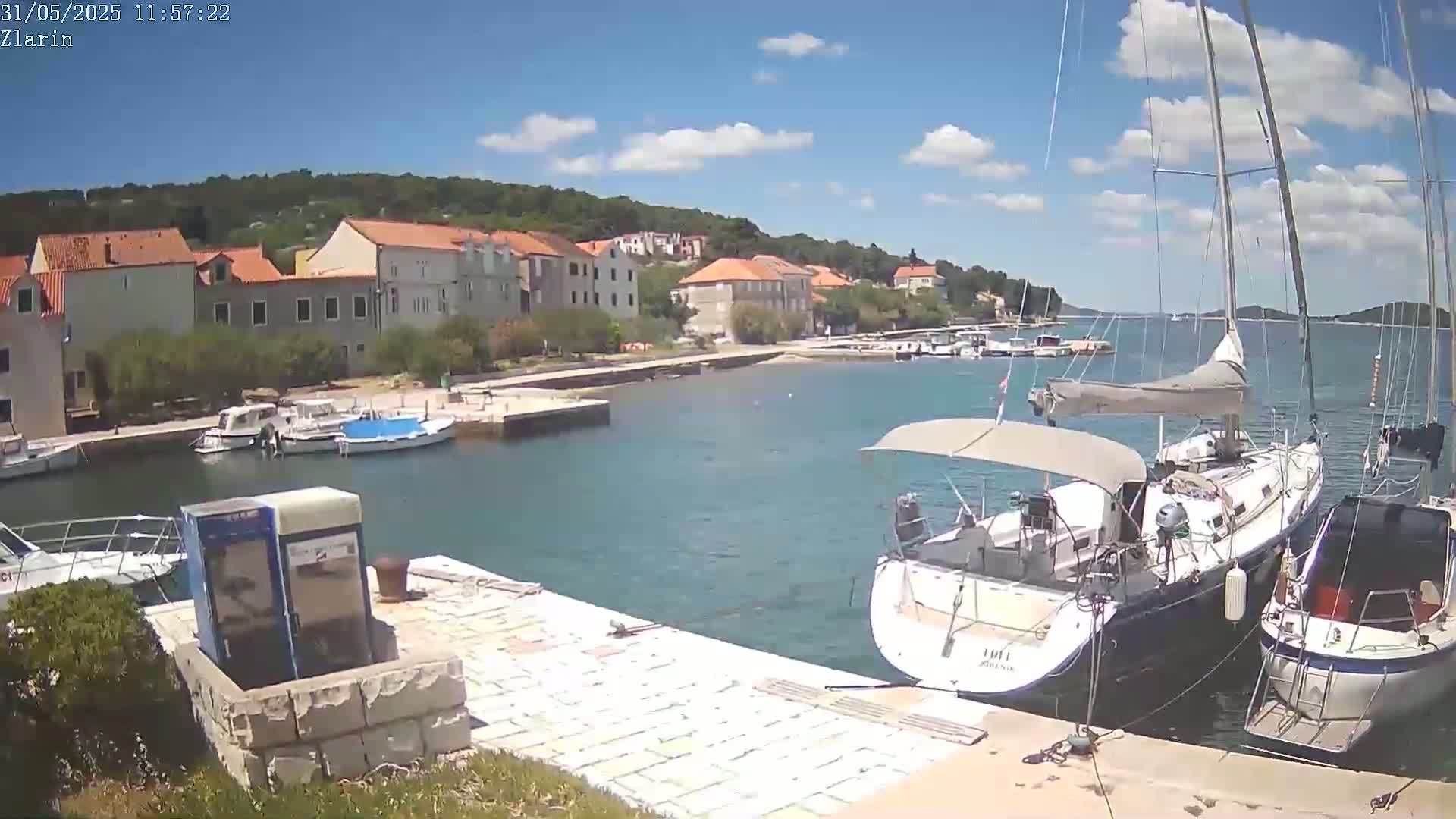 A sunny day shows a harbor with several sailboats and motorboats docked near a row of buildings and green hills.