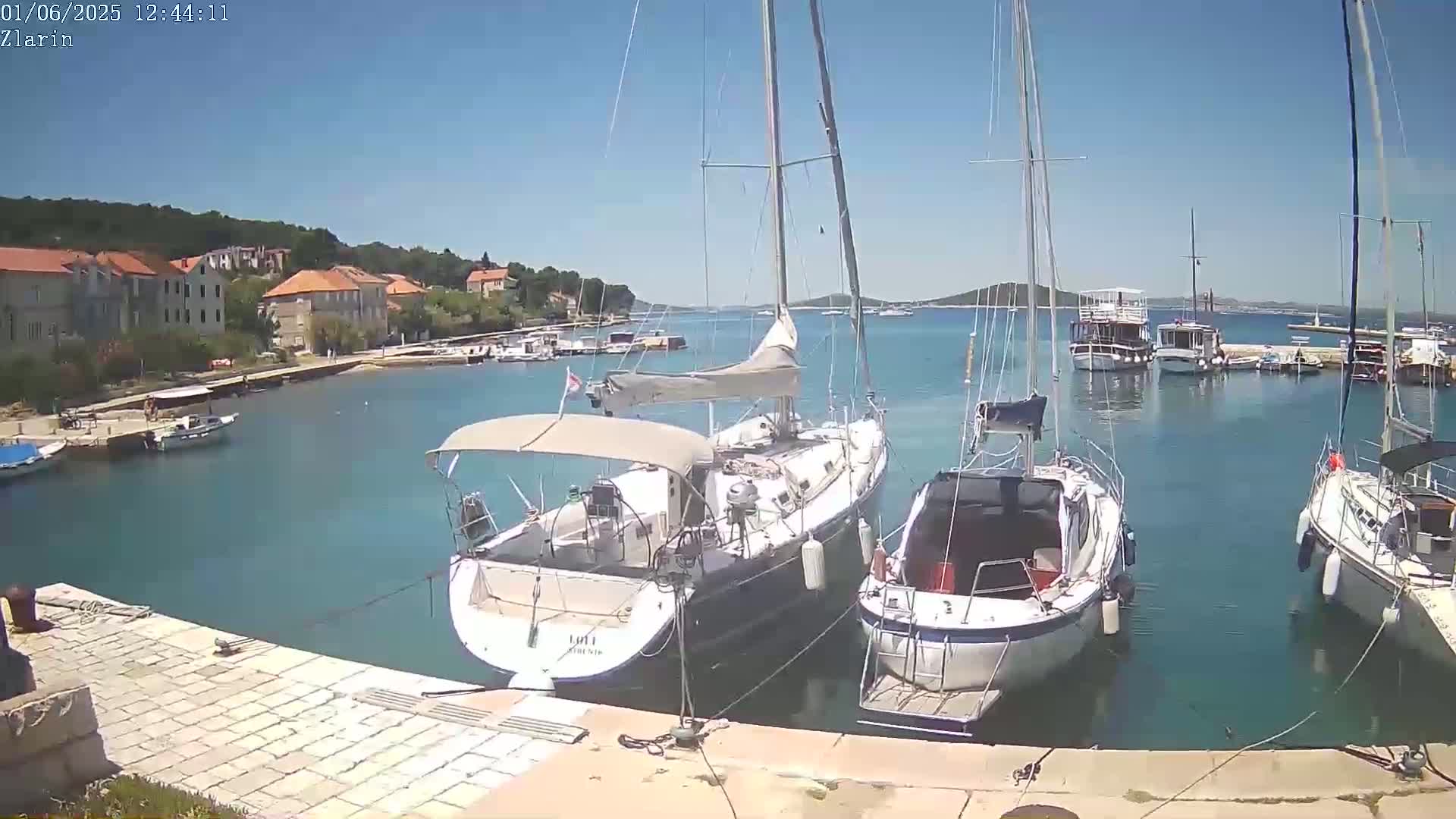 A sunny day at a harbor shows several sailboats and motorboats moored near a stone quay and waterfront buildings.