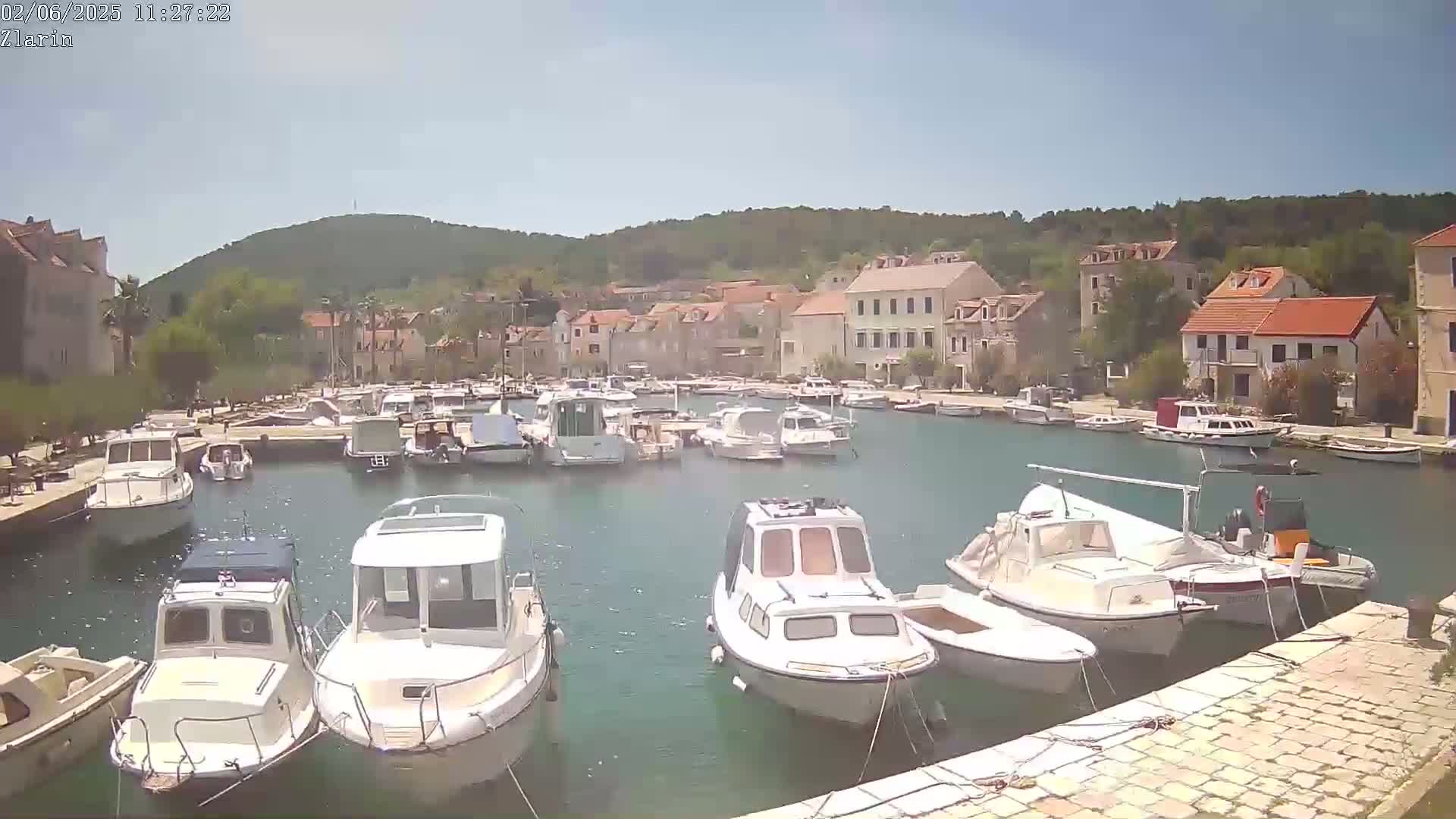A harbor filled with numerous boats is nestled beside a village of stone buildings, all under a clear, sunny sky.
