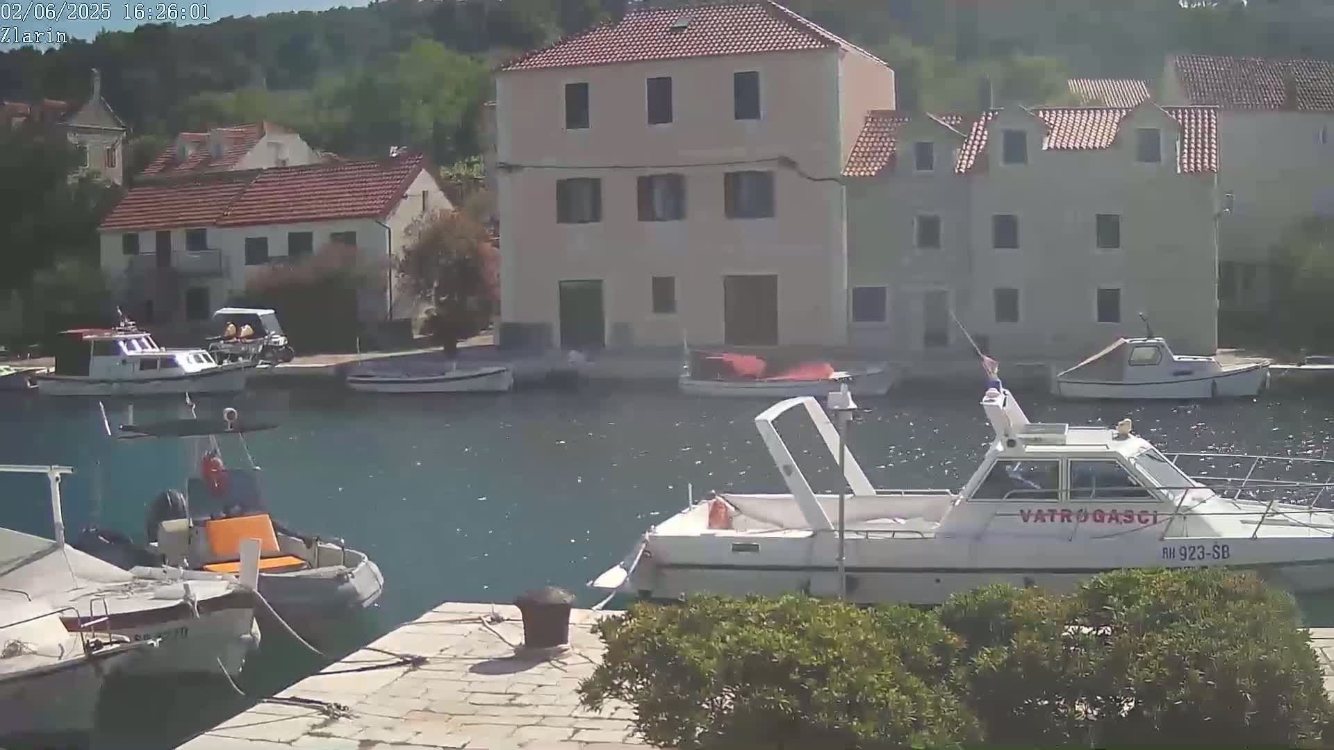 A calm, sunny day shows a harbor with several boats moored, alongside buildings of varying sizes and styles.