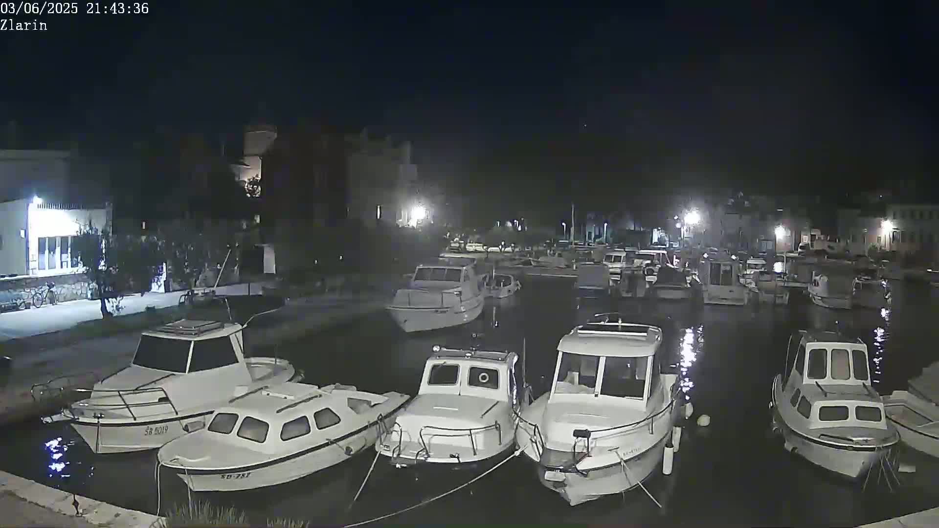 Many small white boats are moored in a calm harbor at night, with buildings visible on the shore.