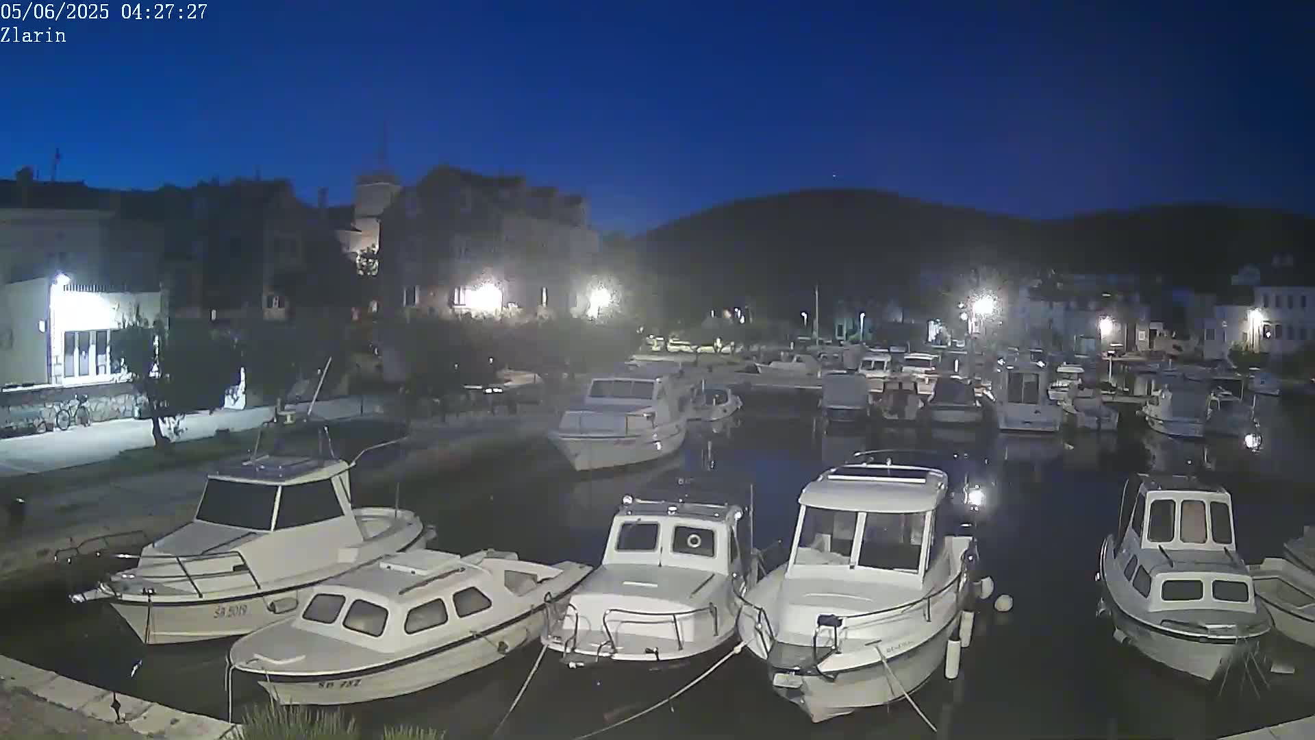 A marina at night under a clear, dark blue sky, filled with numerous small motorboats docked alongside a waterfront lined with buildings.