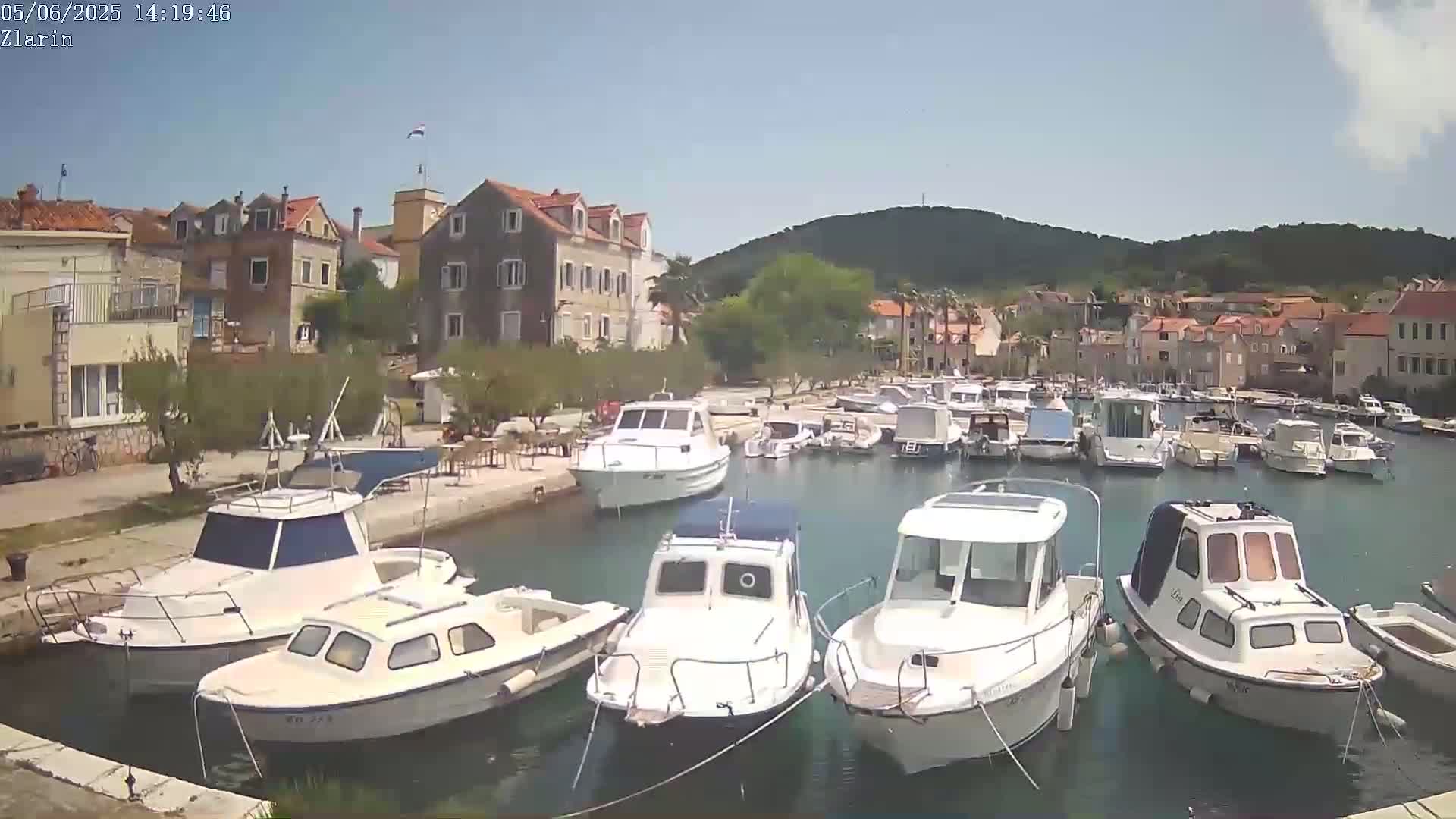 A sunny day shows several white motorboats docked in a calm harbor next to a village of stone buildings and green hills.
