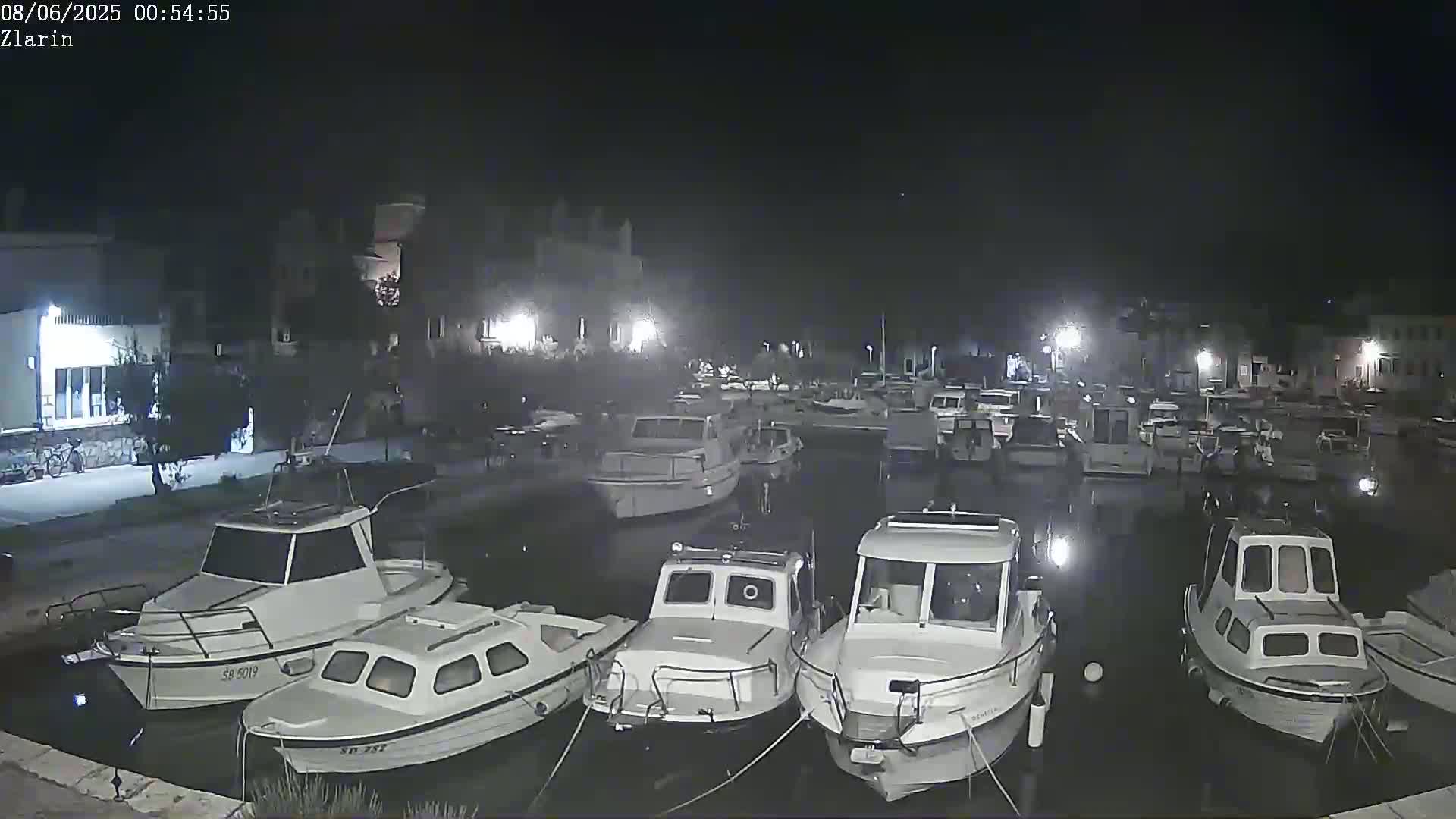 A nighttime view of a marina filled with numerous small white boats moored alongside a stone quay under a dark, clear sky.