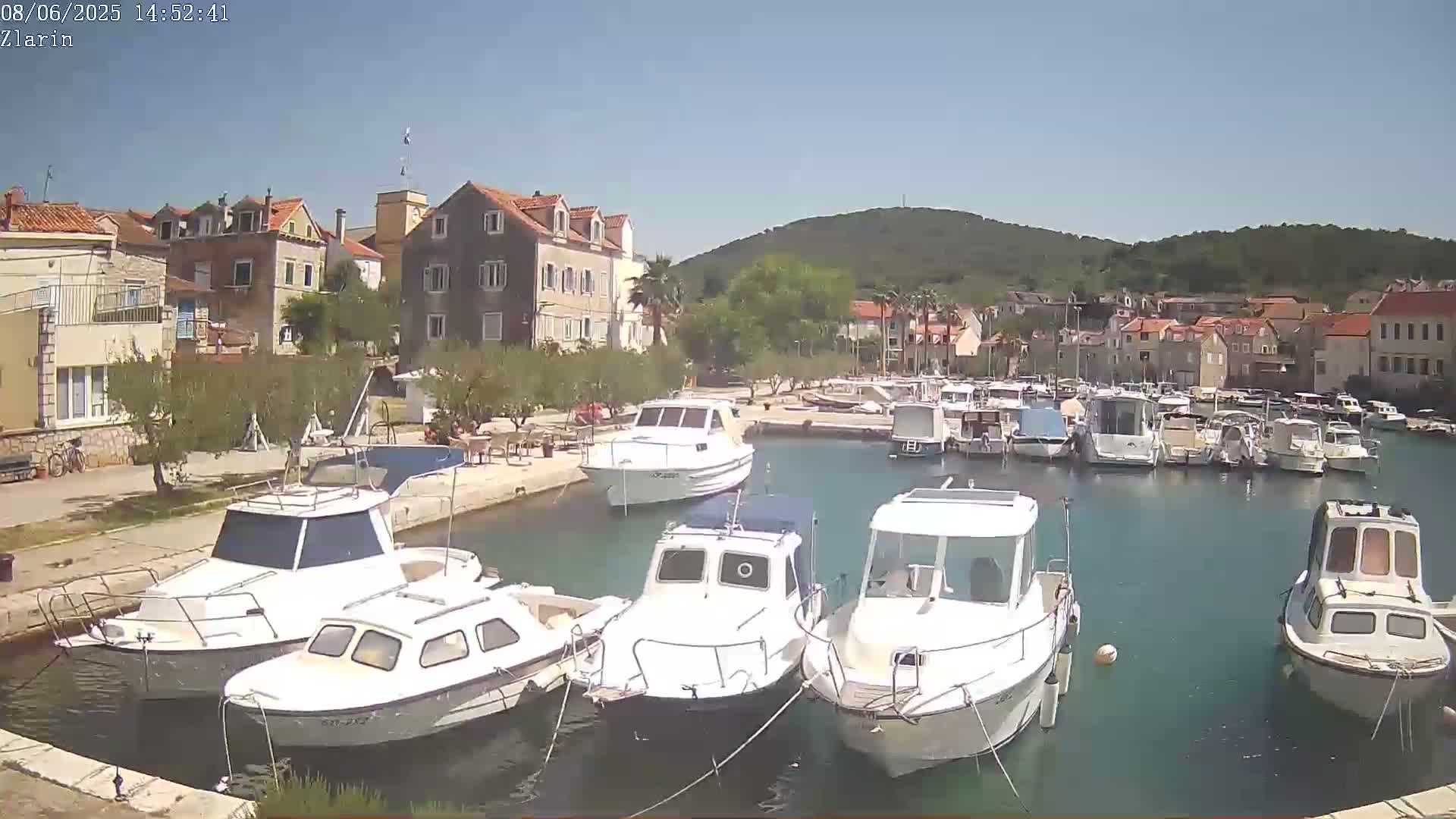 A harbor filled with numerous boats is situated in front of a village of stone buildings under a clear, sunny sky.