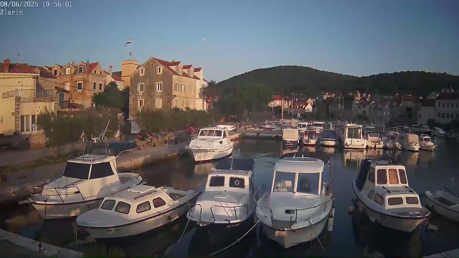 Several white motorboats are moored in a calm harbor next to a stone village under a clear twilight sky.