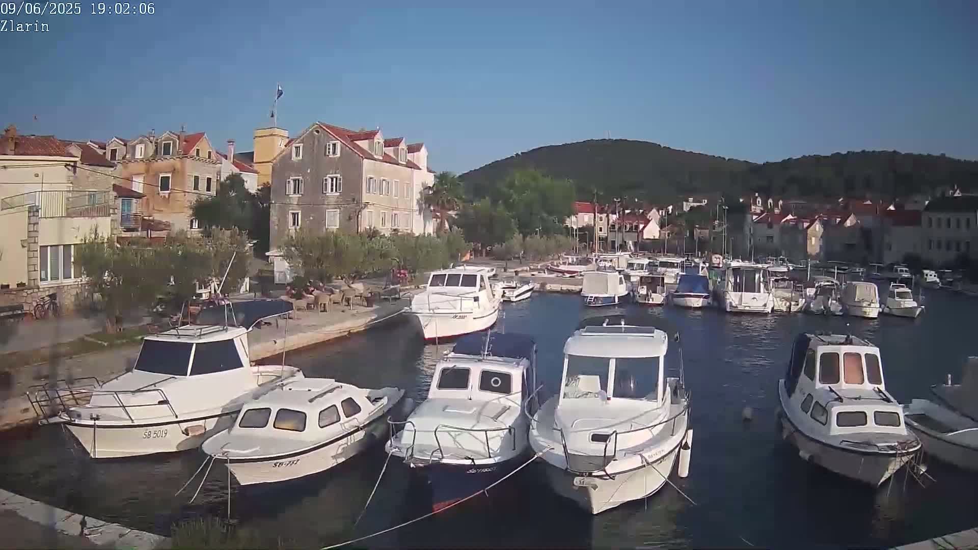 A harbor filled with motorboats is nestled next to a village of stone buildings under a clear blue sky.