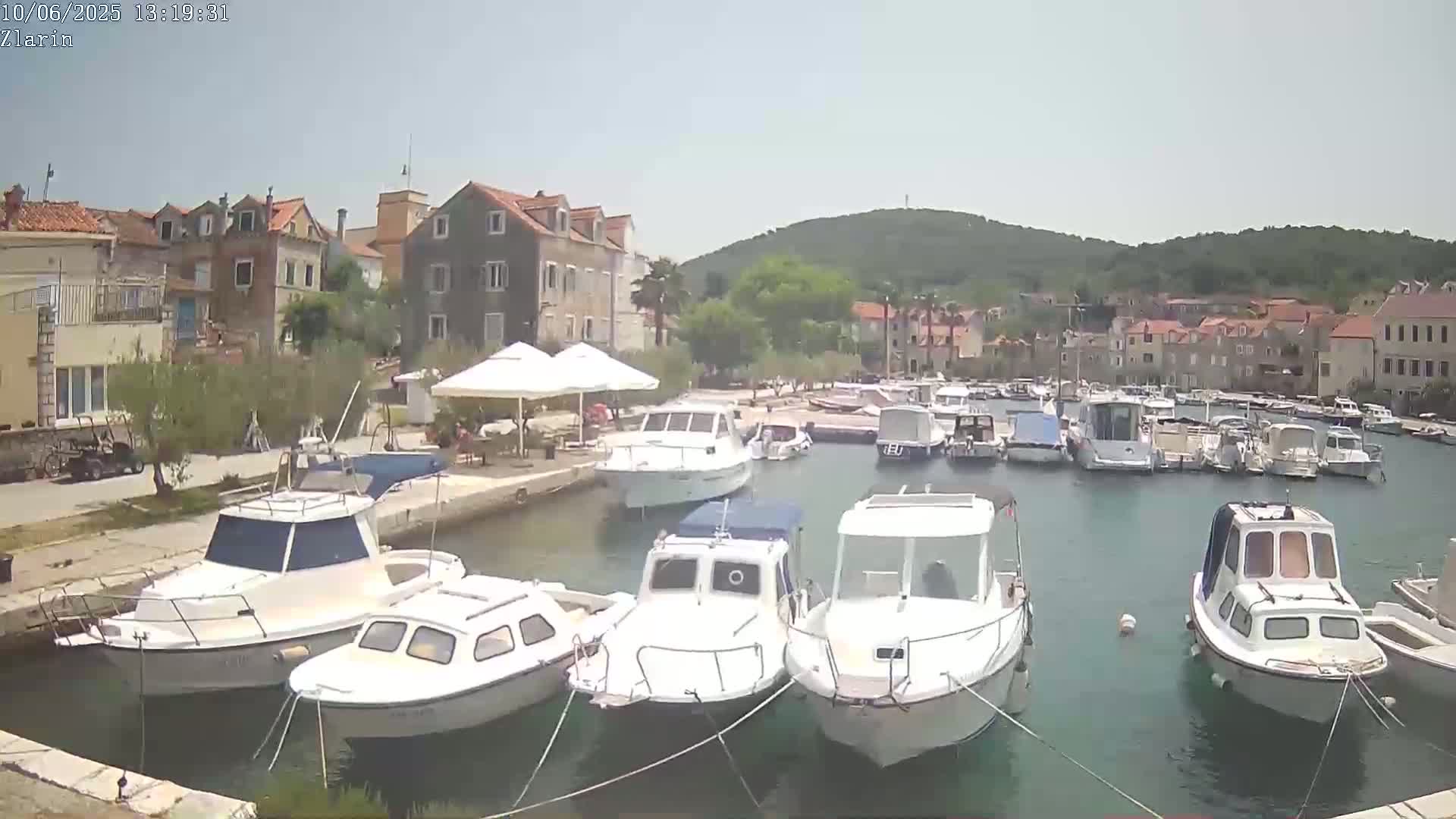 A small harbor filled with numerous white motorboats is situated next to a village of stone buildings under a clear, sunny sky.