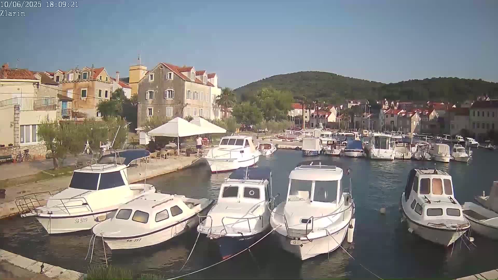 A harbor filled with numerous boats is situated next to a village of stone buildings under a clear, sunny sky.