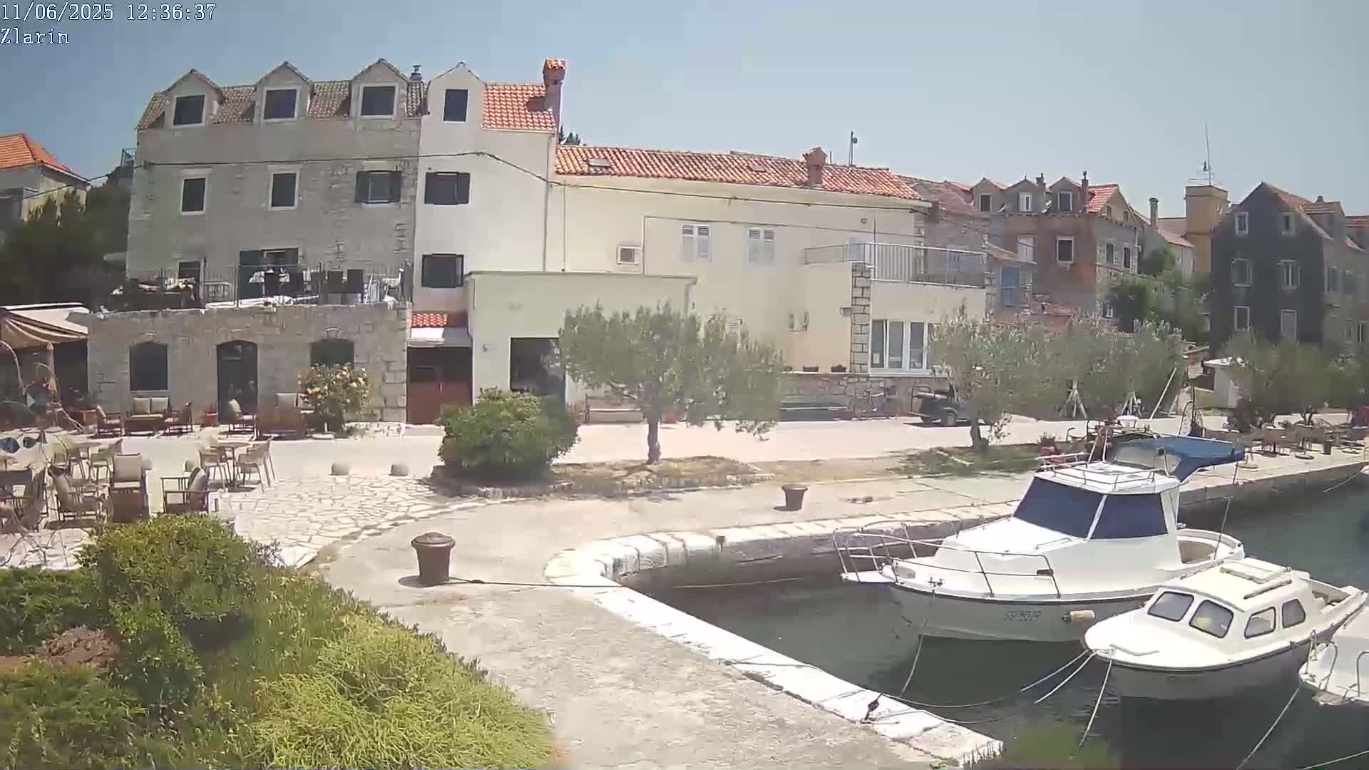 A sunny day reveals a stone building with outdoor seating, a small tree, and two boats moored in a harbor.