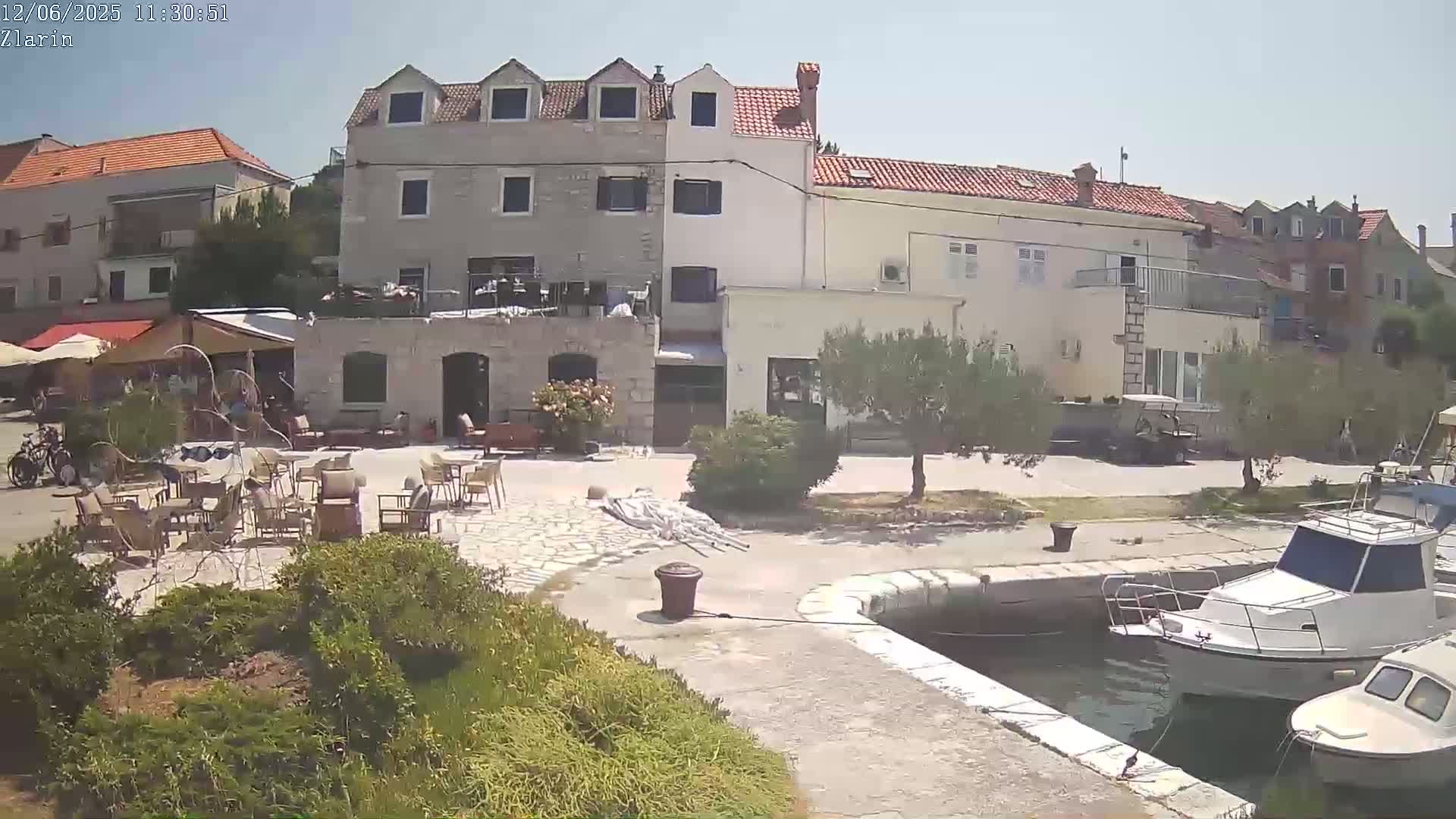 Under a sunny sky, a waterfront plaza with outdoor seating, stone buildings, and moored boats is shown.