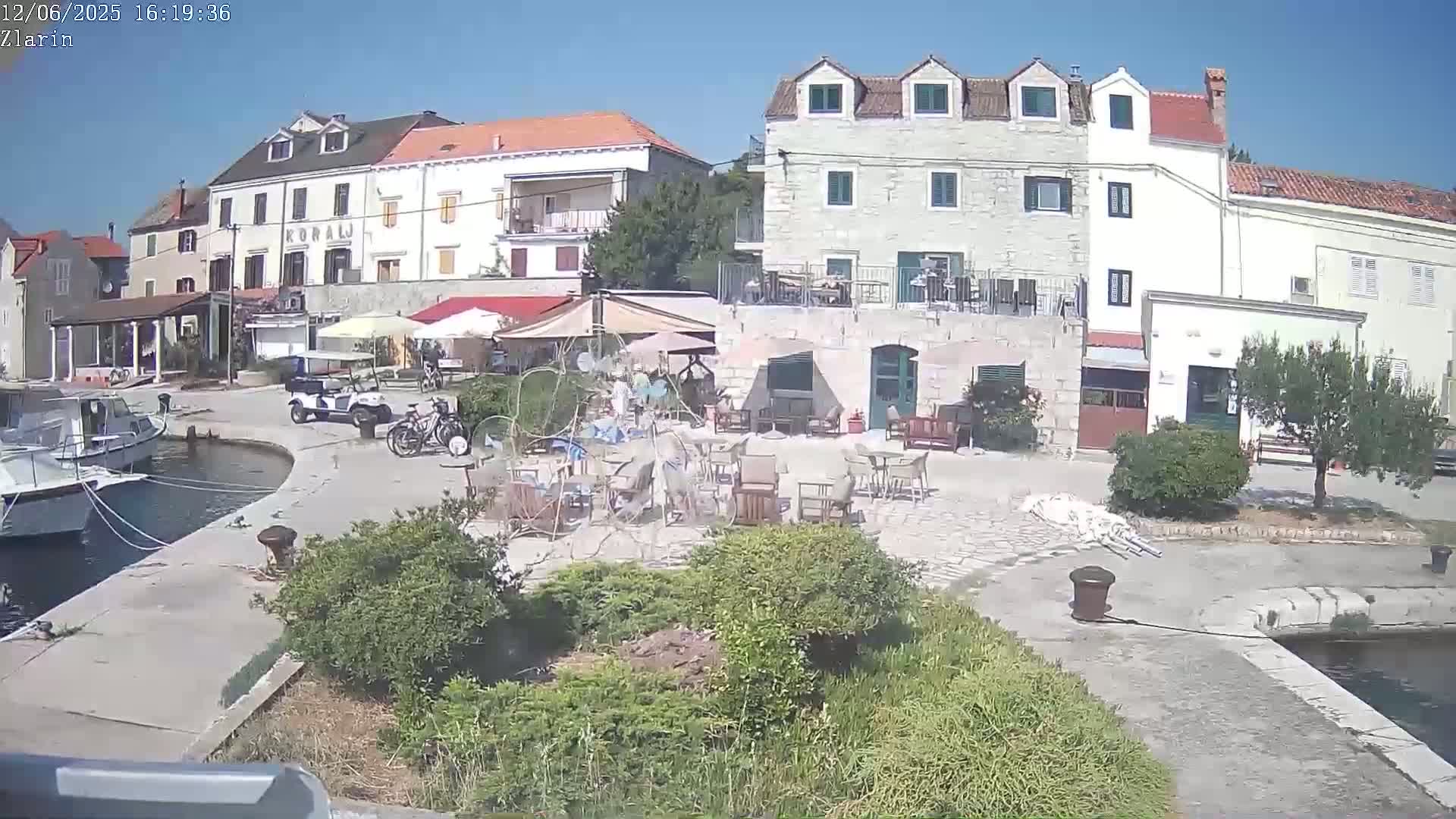 A sunny harbor scene shows stone buildings, outdoor seating areas with tables and chairs, boats moored at a quay, and some vegetation.