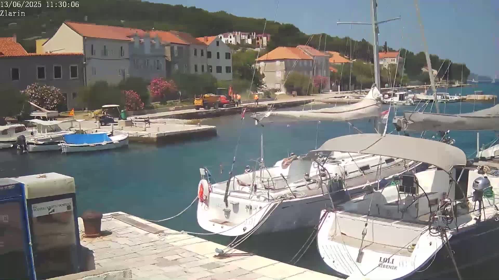 Several sailboats are docked in a harbor next to a village of stone buildings under a sunny sky.