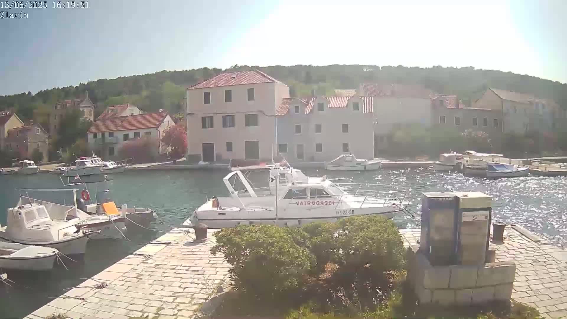 A sunny day shows a harbor filled with several boats, including a fireboat, docked near a small village nestled against a hillside.