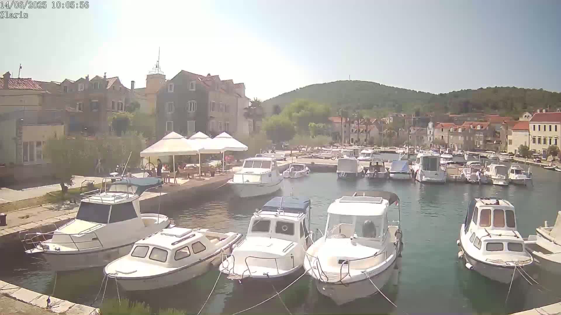 A harbor filled with numerous boats is situated in front of a town on a sunny day, with a hill visible in the background.