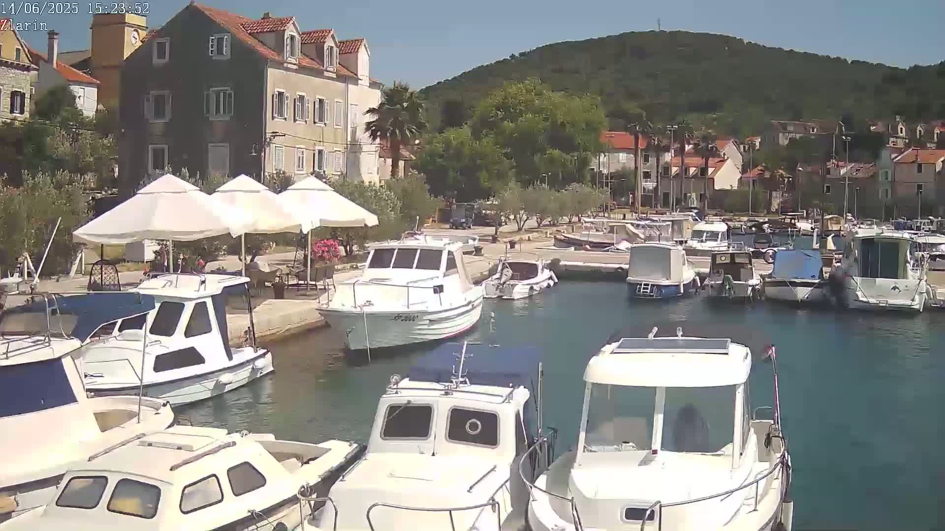 A sunny harbor scene shows numerous white boats moored near a waterfront lined with buildings and shaded by umbrellas.