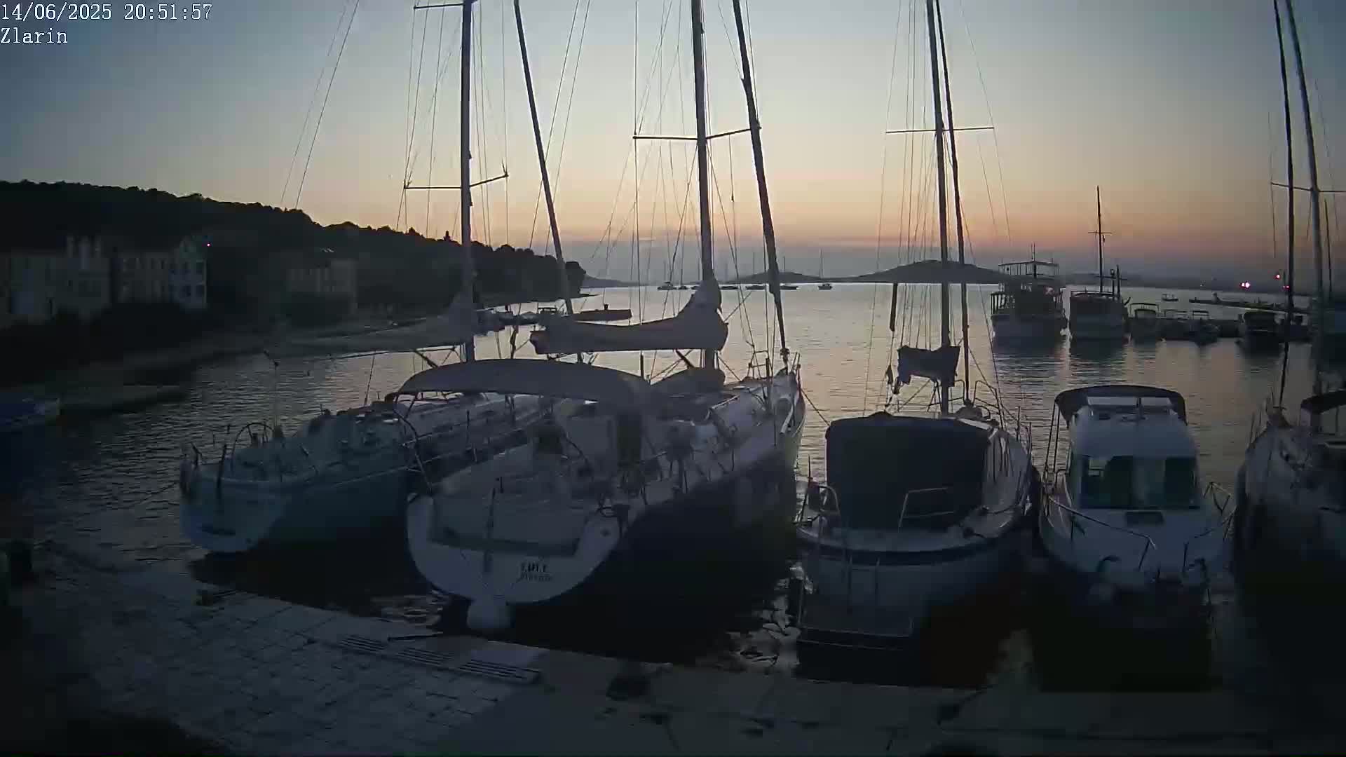 A sunny harbor scene shows numerous white boats moored near a waterfront lined with buildings and shaded by umbrellas.
