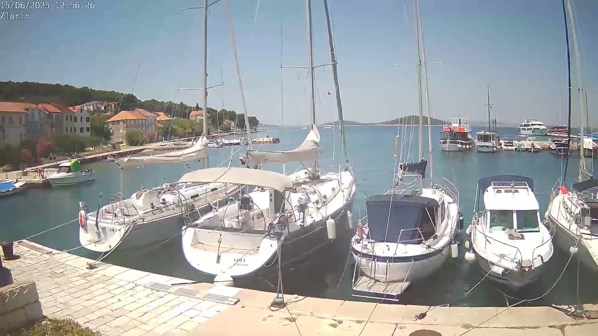 A sunny day shows several sailboats and motorboats moored in a harbor next to a stone waterfront and buildings.