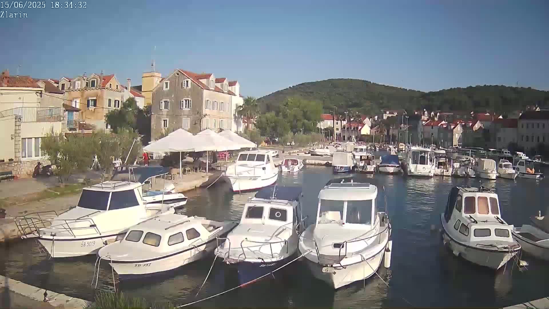 A harbor filled with numerous boats is nestled beside a village of stone buildings under a clear, sunny sky.