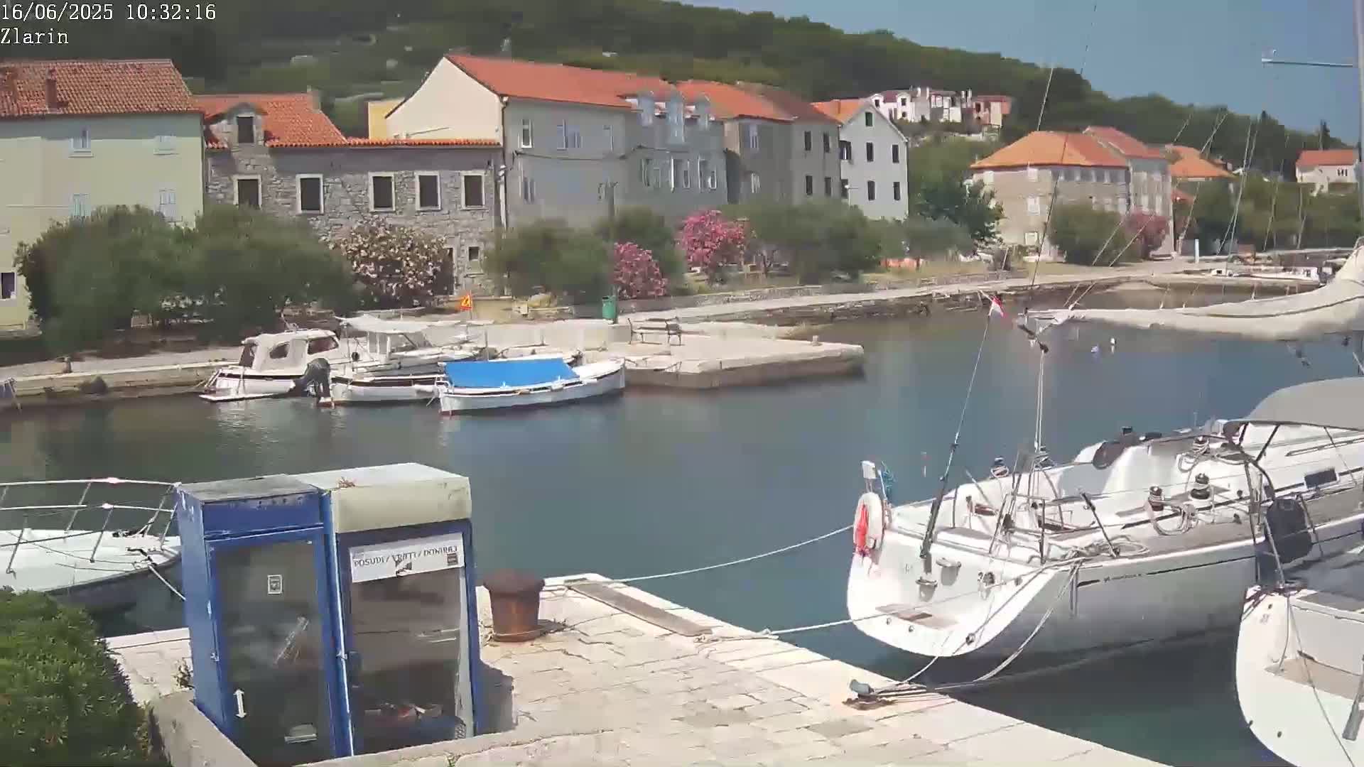 A sunny day at a harbor shows several small boats docked near a quaint village, with a larger sailboat in the foreground.